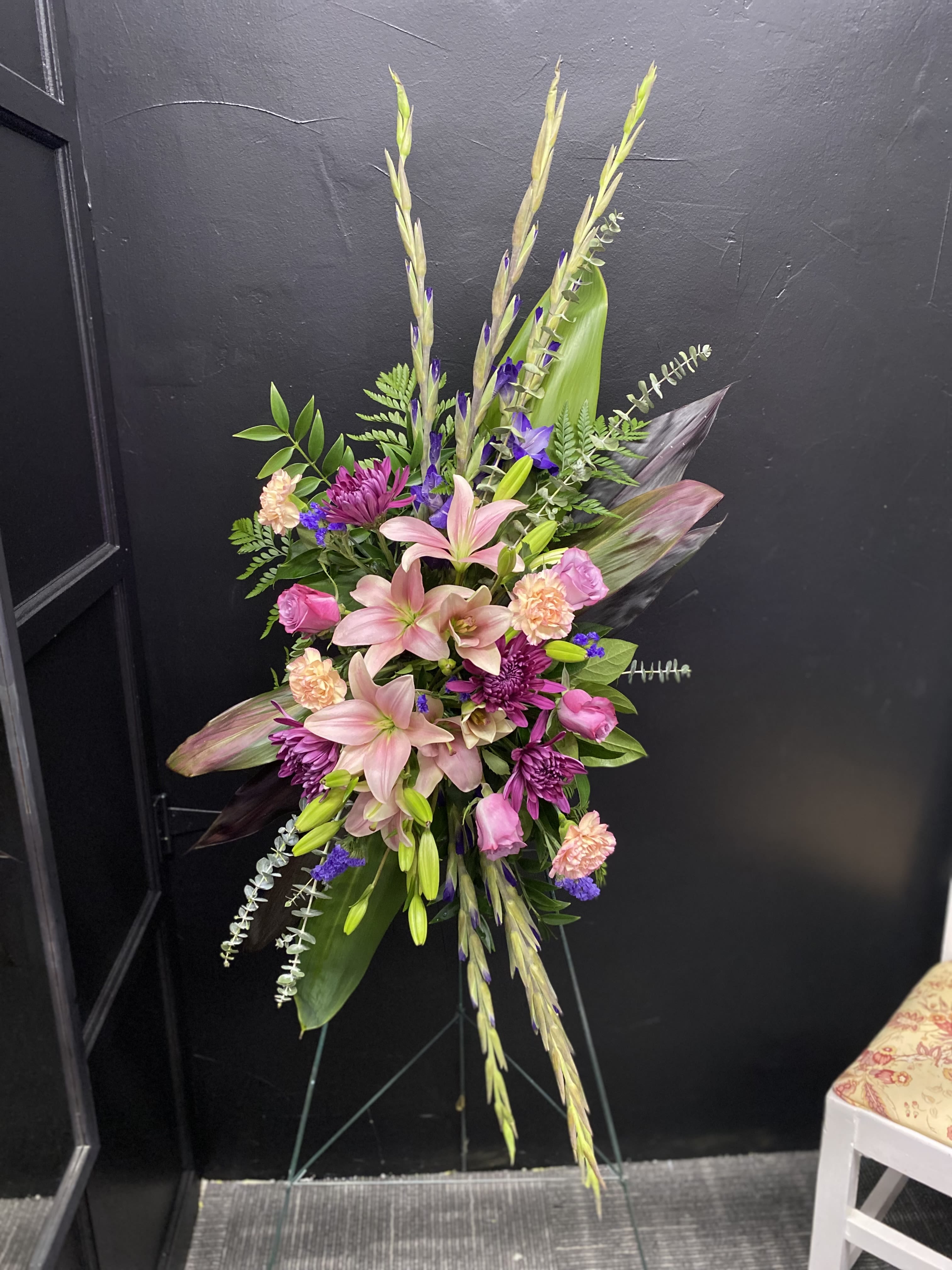 Meadow Song - A tall, layered standing spray that reads as both elegant and deeply expressive-perfect as a sympathy arrangement or for formal occasions like a wedding or anniversary. Pale pink lilies form the striking focal point, their large open blooms giving the piece a soft, fragrant center while magenta and purple chrysanthemums and roses provide bold contrast so the person you're honoring feels genuinely seen.  This arrangement mixes peach carnations and small accent blooms for texture, with long upright grass-like stems and broad tropical leaves to create dramatic height and a graceful, asymmetrical silhouette. Displayed on a metal easel, the piece stands roughly life-size for funeral home or ceremony presentation and carries a refined, ceremonial presence. For a loved one who deserves a dignified, floral tribute or a commemorative display at a wedding or anniversary, this spray delivers a balanced palette of pink, purple, green, white, and peach that reads both romantic and solemn.