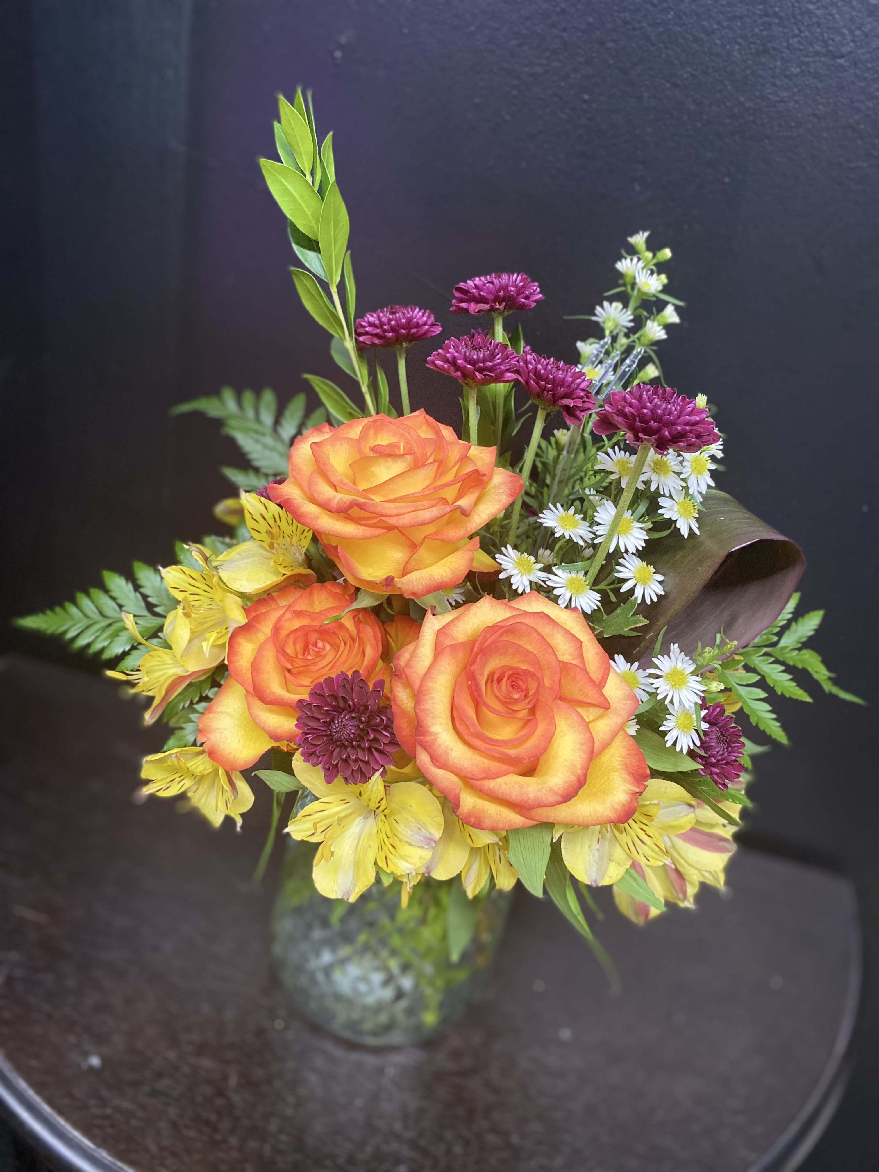 Blooming Bliss - Three vibrant orange roses take center stage in this cheerful bouquet, surrounded by sunny yellow alstroemeria, purple chrysanthemum blooms, and small white aster-like filler flowers to create a lively contrast that feels both festive and thoughtful. Perfect for birthdays, thank-you gifting, or to brighten someone's day, this mixed-flower arrangement reads as joyful and hand-delivered-the kind of bouquet that will make the person you're thinking of feel genuinely celebrated.  Arranged in a clear vase, the design balances casual charm with polished presentation. The mix of textures-the velvety rose petals, delicate alstroemeria blooms, rounded chrysanthemum heads, and dainty white fillers-gives visual depth and a fresh, textured silhouette. Size and stem length work for tabletops and desks; water-ready in the vase, these stems arrive prepared for display.  Send this bright arrangement to mark a cheerful milestone or as a spontaneous pick-me-up; its warm oranges and yellows softened by cool purple accents suit a wide range of recipients from friends to coworkers. For best enjoyment, refresh the vase water every 2-3 days and trim stems slightly when changing water to extend bloom life.
