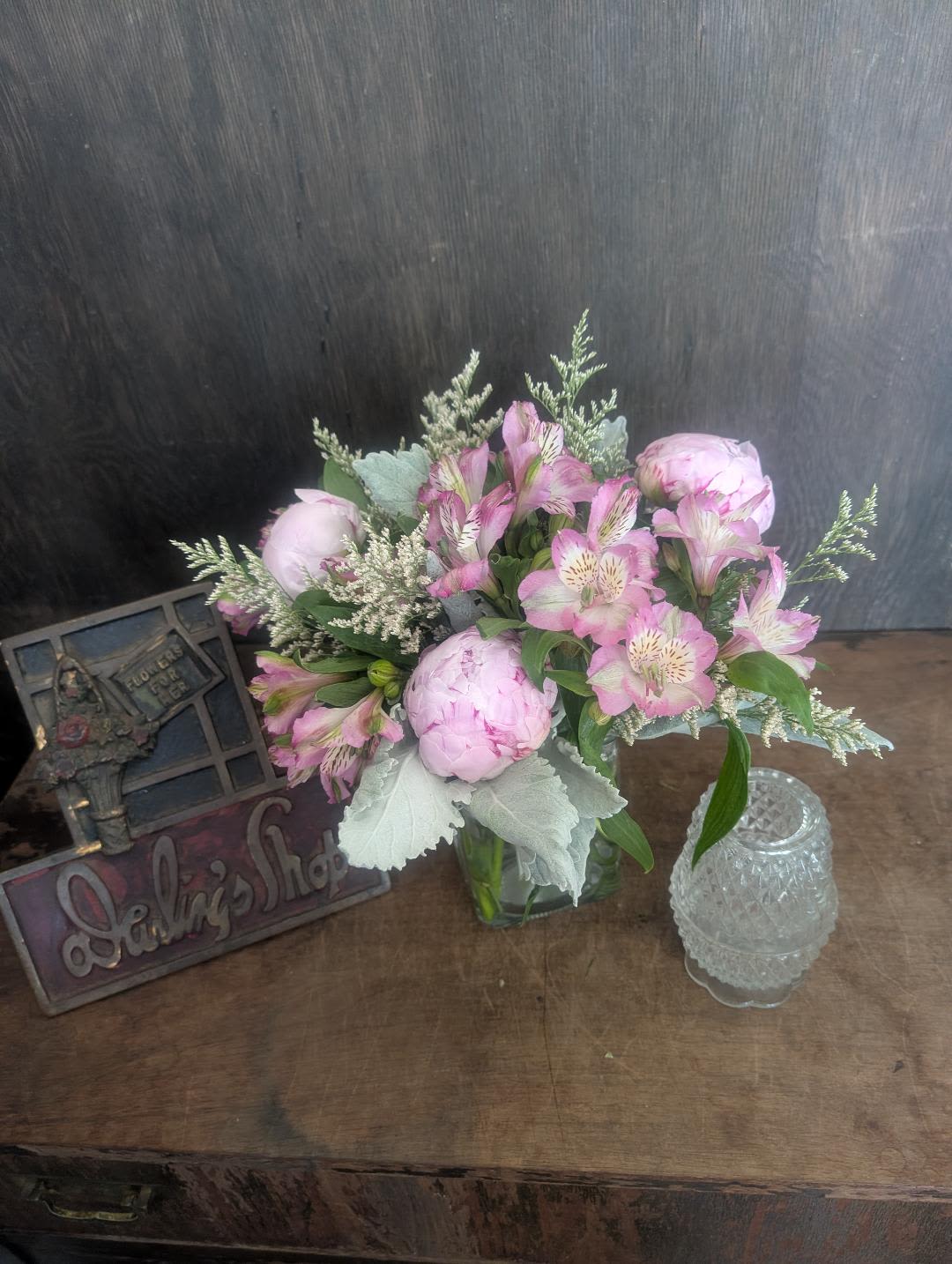 Peonies and Alstroemeria  next day delivery - Soft pink peonies paired with pink-and-white alstroemeria create a romantic, garden-style bouquet that reads like a personal note of care - perfect as an anniversary or birthday surprise, a "thinking of you" pick-me-up, or a gentle housewarming gift. The pastel palette of pink, white, and soft green plus silvery foliage gives the arrangement a serene springtime feel that still works for year-round occasions like Just Because, Get Well, or Mother's Day.  Arranged loose and low in a clear glass vase, the blooms are accented with pale filler sprays for texture and a cool contrast to the lush peony petals. The display is styled for home presentation and comes beautifully complemented by a decorative sign and a glass candle holder to help set a cozy scene. Sized to sit on a dining table or bedside table, the composition brings a soft, fragrant focal point that will make the person you're thinking of feel truly seen and loved.