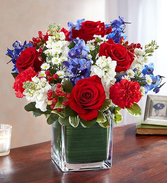 Red roses, blue flowers, and white daisies arranged in a square glass vase