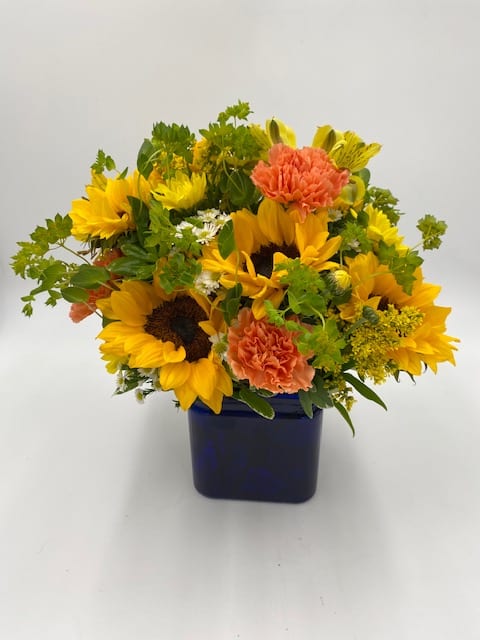 Low arrangement of yellow sunflowers and orange carnations in a blue cube vase