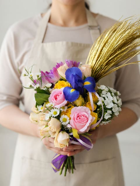 Freestyle Freshies - Soft pink and pale yellow roses mingle with vivid blue irises and cheerful white daisy-like blooms in this handheld flower arrangement-perfect as a birthday bouquet, a springtime gift, or a bright thank-you gesture. The combination of soft pastels and saturated accents gives the recipient an immediate lift: she'll feel noticed and celebrated the moment she sees the playful color mix and unexpected pop of blue.  Hand-tied with purple ribbon and a touch of metallic gold decorative grass for height and sparkle, the Freestyle Freshies pairs classic roses and daisies with modern textural contrast. Sizes are compact and easy to carry, arranged to sit comfortably in the hand or in a small vase; the gold spray adds festive movement without overpowering the blooms.  Choose this bouquet when you want a cheerful, slightly whimsical arrangement that reads as both modern and heartfelt-ideal for birthdays, congratulations, spring events, or simply saying "I'm thinking of you." Standard care: trim stems, change water daily, and remove wilted foliage to keep the mixed blooms fresh longer.