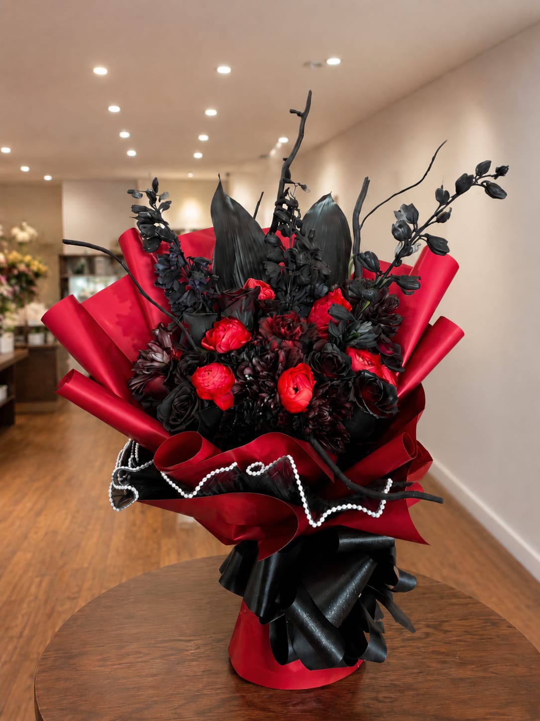 Midnight Passion Bouquet - A sophisticated, modern floral arrangement in shades of deep red and rich black, designed to convey luxury, passion, and elegance. Red ranunculus stand out against dark, velvety roses, black foliage, and artistic accents that add height and drama. Wrapped in red and black paper with an elegant finish and decorative pearls, it creates an exclusive presentation—perfect for surprising someone on special occasions, anniversaries, or romantic declarations, or for making a high-impact statement.