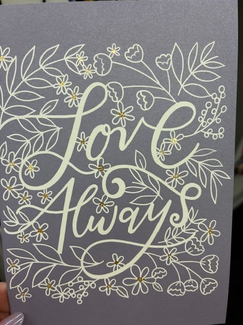 Love Always card - A delicate lavender greeting card that says "Love Always" in flowing script creates a soft, romantic moment for anniversaries, weddings, or just because. The pale purple background and white floral line art feel intimate and thoughtful-the small yellow accents at each drawn flower center add a sunny, optimistic pop that makes the recipient feel seen and cherished. This card reads as gentle and sincere, perfect for expressing love, gratitude, or a quiet "thinking of you."  Inside, the card's restrained design keeps the message front and center: a tasteful, sentimental choice for thank-you notes, romantic messages, or a simple everyday gesture of care. Pair it with yellow, white, or purple blooms to echo the palette and create a coordinated gift; the card's size and fold make it easy to tuck into an arrangement or attach to a bouquet. Whether marking Valentine's Day, an anniversary, or sending comfort and appreciation, this card helps your person feel truly loved.