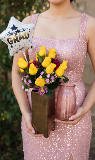 Mini, Single balloon,balloon bunch - Bright yellow roses take center stage in this cheerful flower arrangement, paired with soft pink and purple daisy-like chrysanthemums for a fresh, celebratory look-perfect as a graduation bouquet, birthday surprise, or "just because" pick-me-up. The included star-shaped foil balloon reading "Congrats Grad" makes the gift instantly recognizable for commencement, while the playful round and cluster foil balloons work equally well for birthdays, new baby celebrations, anniversaries, or a thoughtful Thinking of You moment.  Arranged in a tall bronze-toned metal vase and paired with a glass candle holder for an elevated presentation, the mix of vivid yellow, gentle pink, and lavender tones creates lively contrast and a youthful, upbeat texture. Roses and mums are arranged to show off the blooms' color and form; the glossy foil balloons add height and sparkle so the recipient feels celebrated the moment it arrives.  Send this as a graduation centerpiece, congratulations gift, anniversary surprise, or welcome-baby bouquet-the combination of flowers, metallic vase, and festive balloons reads as both party-ready and keepsake-worthy. Care: keep flowers in fresh water, away from direct heat, and gently re-cut stems every few days to extend bloom life.