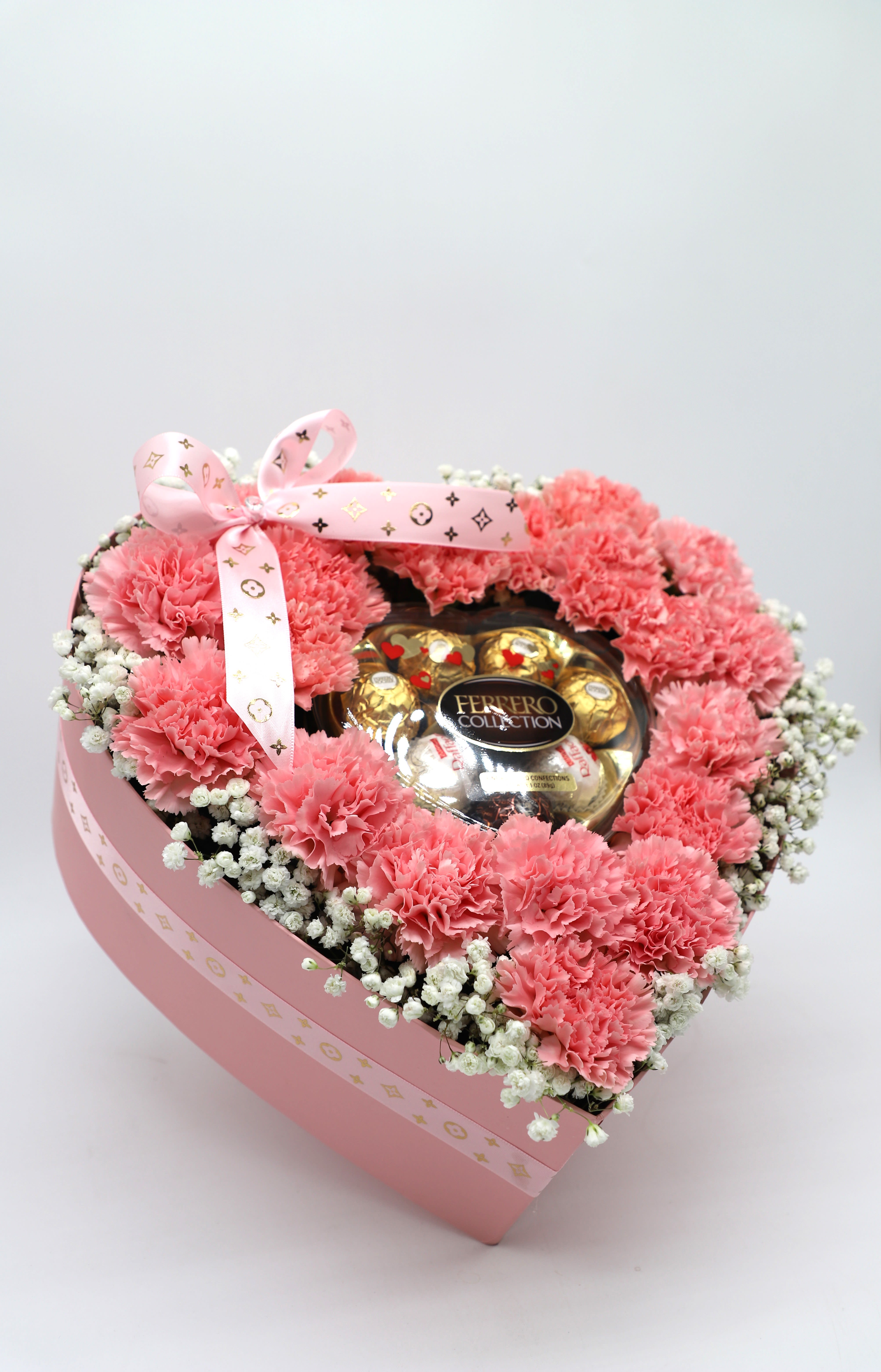 Pink carnations arranged around a Ferrero Rocher chocolate box in a pink heart-shaped box