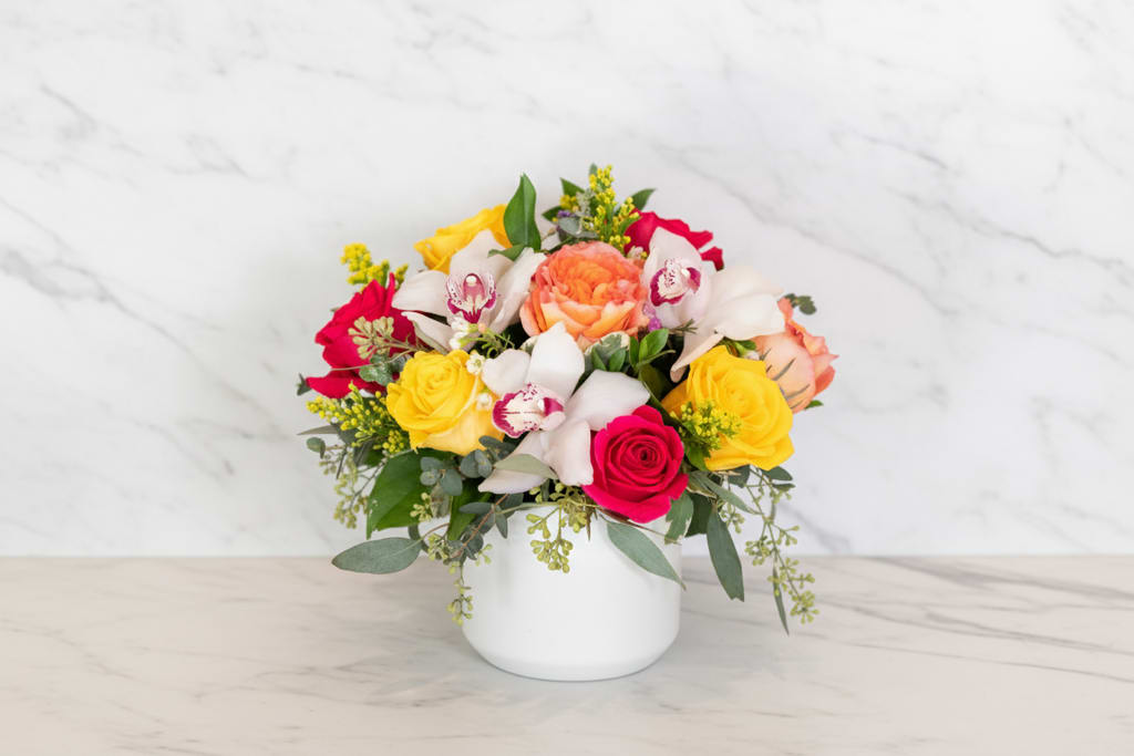 Summer on Polk Street - A compact, cheerful bouquet that reads like summertime in bloom-Summer on Polk Street is a mixed-color flower arrangement perfect for a birthday bouquet, thank-you gift, or a bright pick-me-up for someone recovering or celebrating. The rounded design pairs pink and yellow roses with white orchids and a peach-toned focal bloom to create a warm, joyful impression the recipient will notice the moment it arrives.  This arrangement includes 9 roses complemented by orchids, small yellow filler flowers, and eucalyptus-like greenery for texture and softness. Flowers are arranged in a clean white ceramic vase that keeps the look modern and polished while letting the saturated pinks, yellows, whites, and soft peach tones take center stage. Specific rose varieties and the exact vessel may vary with seasonal availability.  Ideal for summer celebrations but equally at home for Mother's Day, anniversaries, graduations, or simply saying "I'm thinking of you," this versatile design sends an unmistakable message of warmth and care. Order with confidence for birthday deliveries, get well wishes, congratulations, or a thoughtful just-because surprise; care is simple-fresh water and a cool spot will keep the arrangement looking its best.