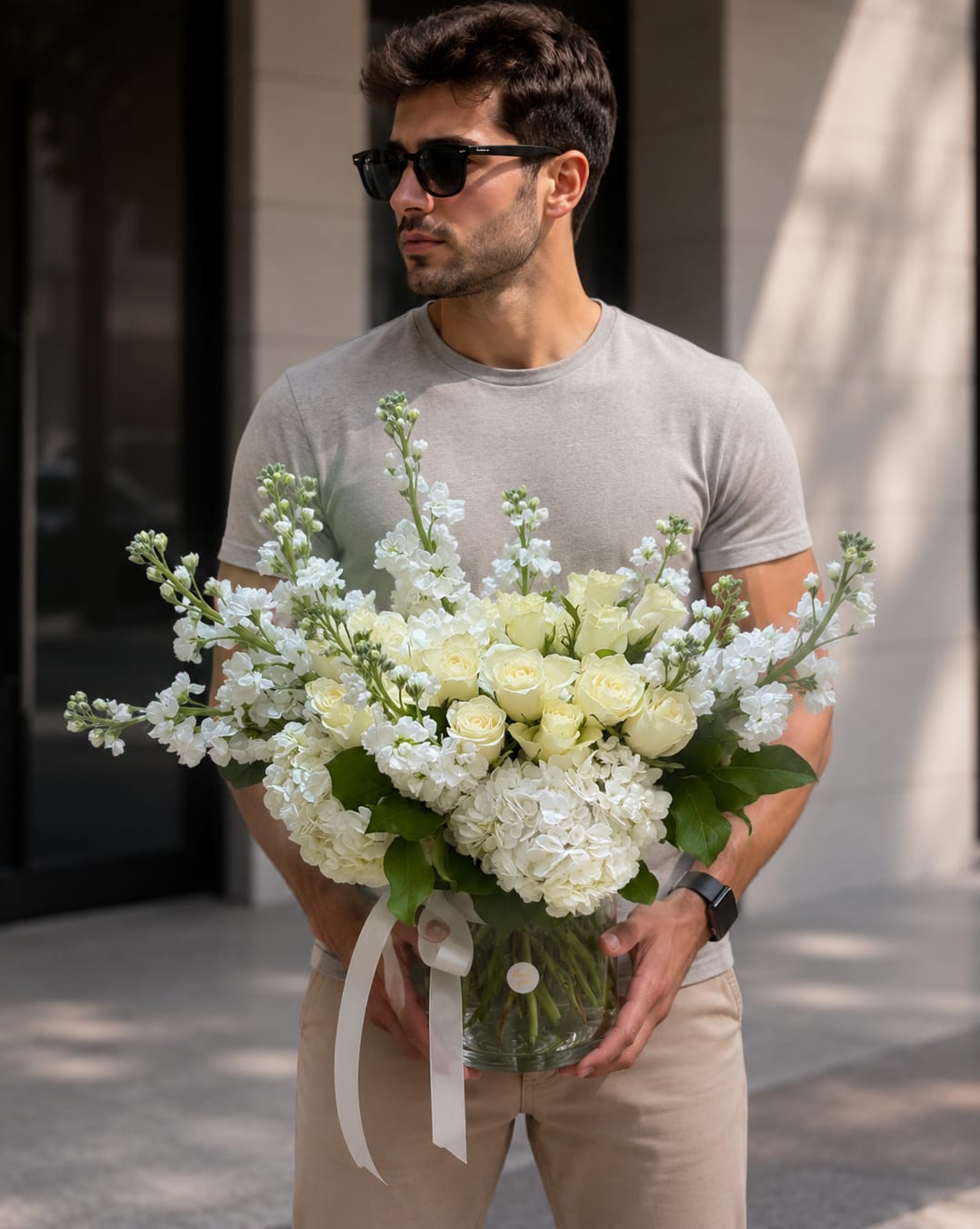 Compliment Lux  - Creamy white roses and full white hydrangeas form a serene, rounded bouquet that says "I'm sorry" with quiet elegance - a sympathy or thinking-of-you flower arrangement that also works beautifully for thank-you gestures. Tall white stock adds soft, airy height while pale yellow accents and fresh green leaves give the composition a gentle warmth that feels comforting and sincere.  Arranged low and full in a clear glass vase and tied with a long white ribbon, the design balances lush, textured hydrangea heads with the refined shape of roses and upright stock. The palette of white, ivory, pale yellow, and green keeps the look classic and versatile for sympathy and funeral services, a thoughtful thank-you, or a tender Mother's Day or Parent's Day gift. Size and exact stem counts vary by season; stems are arranged to showcase fullness and movement and arrive ready to display with fresh water in the vase.