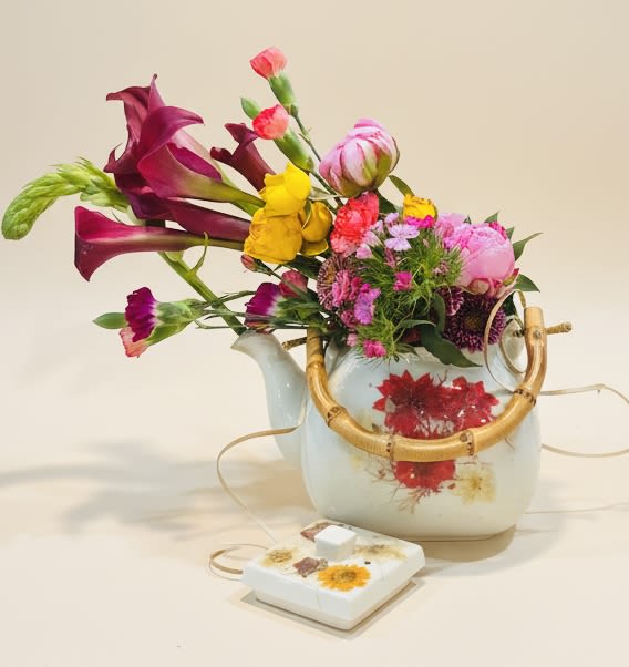 The Tailored Teapot (flower andesin designed) - Deep magenta calla lilies arching over sunny yellow blooms make a bold first impression in this anniversary and birthday-ready teapot arrangement - the Tailored Teapot is a flower arrangement that reads like a cheerful, handcrafted gift. Designed to celebrate love and everyday joy, it brings a lively, feminine mix of color and texture that feels equally right for Romance, Just Because surprises, Get Well wishes, or a thoughtful Housewarming present.  Inside a white ceramic teapot vase with a bamboo handle, bright yellow flowers contrast with saturated purple calla lilies, soft pink carnations, delicate peony touches, and small daisy-like blooms for a compact, asymmetrical profile that seems to spill outward. A decorative ribbon and a small matching box finish the presentation, making the set feel ready to gift. The size is intimate enough for tabletops and bedside stands but bold enough to make someone feel truly celebrated.  Perfect for saying Thank You, Thinking of You, Retirement congratulations, or seasonal moments like Mother's Day and Persian New Year, this arrangement is crafted to arrive fresh and fragrant. Care is simple: keep flowers in a cool spot away from direct sun, refresh water and trim stems as needed to extend vase life - an effortless, beautiful way to brighten someone's day.