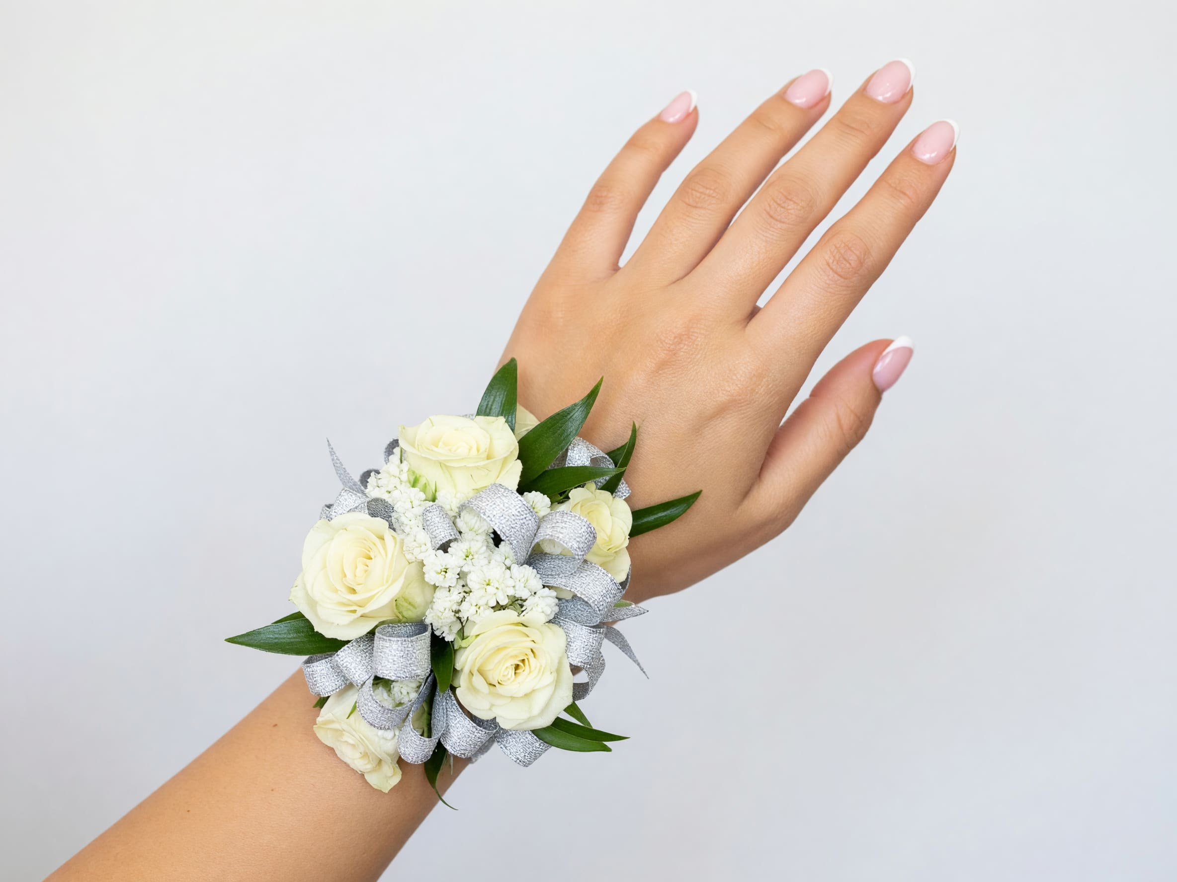 White Mini Rose wrist corsage - A delicate cluster of white mini roses arranged as a wrist corsage creates an elegant finishing touch for weddings, proms, or other dressy events. This white mini rose wrist corsage is designed to make the person you're gifting feel polished and put-together-soft white blooms framed by glossy green leaves read as timeless and refined for a bride, bridesmaid, or honored guest.  The compact design features small white roses paired with clustered white filler flowers for texture, all tucked into a comfortable wristband-style base. A metallic silver ribbon is woven through the arrangement to add a formal, polished sheen and subtle contrast against the fresh green foliage. The corsage's mixed white-and-green palette keeps the look classic while the silver detail gives it a modern, dressy edge.  Perfect for wedding parties, prom dates, mothers of the couple, or anyone who prefers a subtle, sophisticated accessory, this wrist corsage slides on easily and stays close to the wrist for all-day wear. Treat it as a finishing touch that helps the recipient feel truly celebrated; order timing and size options are available at checkout to ensure peak freshness for your event.