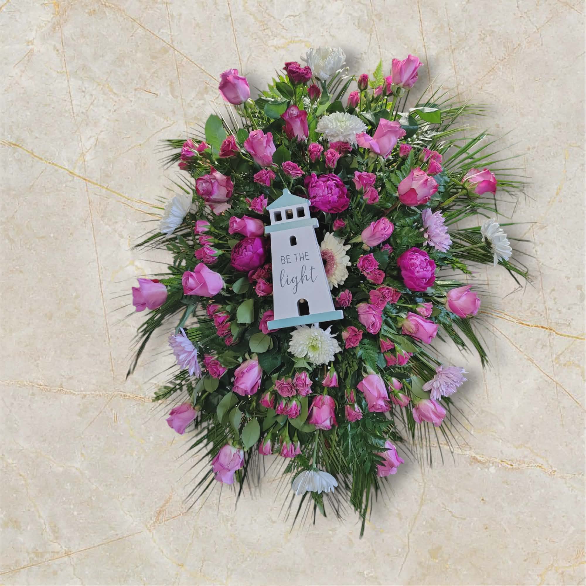Guiding Light - A full standing spray of soft pink roses and small pink spray blooms creates a comforting, hopeful focal point-perfect as a sympathy or funeral flower arrangement to honor someone who guided others with warmth. The arrangement's gentle white daisy-like blossoms and mixed greenery give it a bright, uplifting presence that reads as both romantic and reverent; the decorative lighthouse sign at the center, inscribed "Be the light," offers a quiet, inspirational message for the person you're remembering.  Layered palm-like fronds and airy greenery extend beyond the spray’s edge, adding movement and a natural silhouette that looks beautiful .The combination of roses, chrysanthemums, and daisies in pink, white, and green provides soft color contrast and varied textures-velvety petals, cheerful centers, and feathery foliage-for a thoughtful tribute that comforts and celebrates the recipient's guiding spirit.