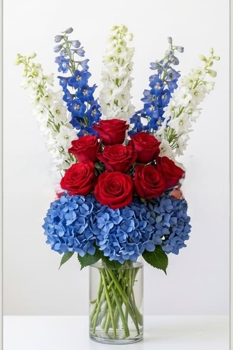 The Stars and Stripes - USA - Bold red roses take center stage in this floral tribute to team spirit-a spirited flower arrangement designed for the FIFA World Cup collection and perfect for game-day cheering, patriotic celebrations, or congratulating a devoted fan. The combination of red, blue, and white delivers an unmistakable national flair: it reads loud and proud while still feeling refined enough for a birthday, victory party, or special recognition.  A dense band of blue hydrangeas anchors the base, giving the design a lush, rounded silhouette, while tall blue and white floral spikes rise behind the roses to add movement and a dramatic vertical outline. The mix of roses, hydrangeas, and complementary seasonal blooms creates rich texture and depth against bright green foliage. Arranged in a clear glass vase, the long stems are visible for a modern, elegant presentation that keeps the focus on color and form.  Give this to the soccer fan who lives for the match-the kind of bouquet that makes them feel celebrated and seen. The designer has curated each bloom for balanced impact and lasting presence; place in fresh water on arrival and refresh water every two days to extend the life of the flowers.