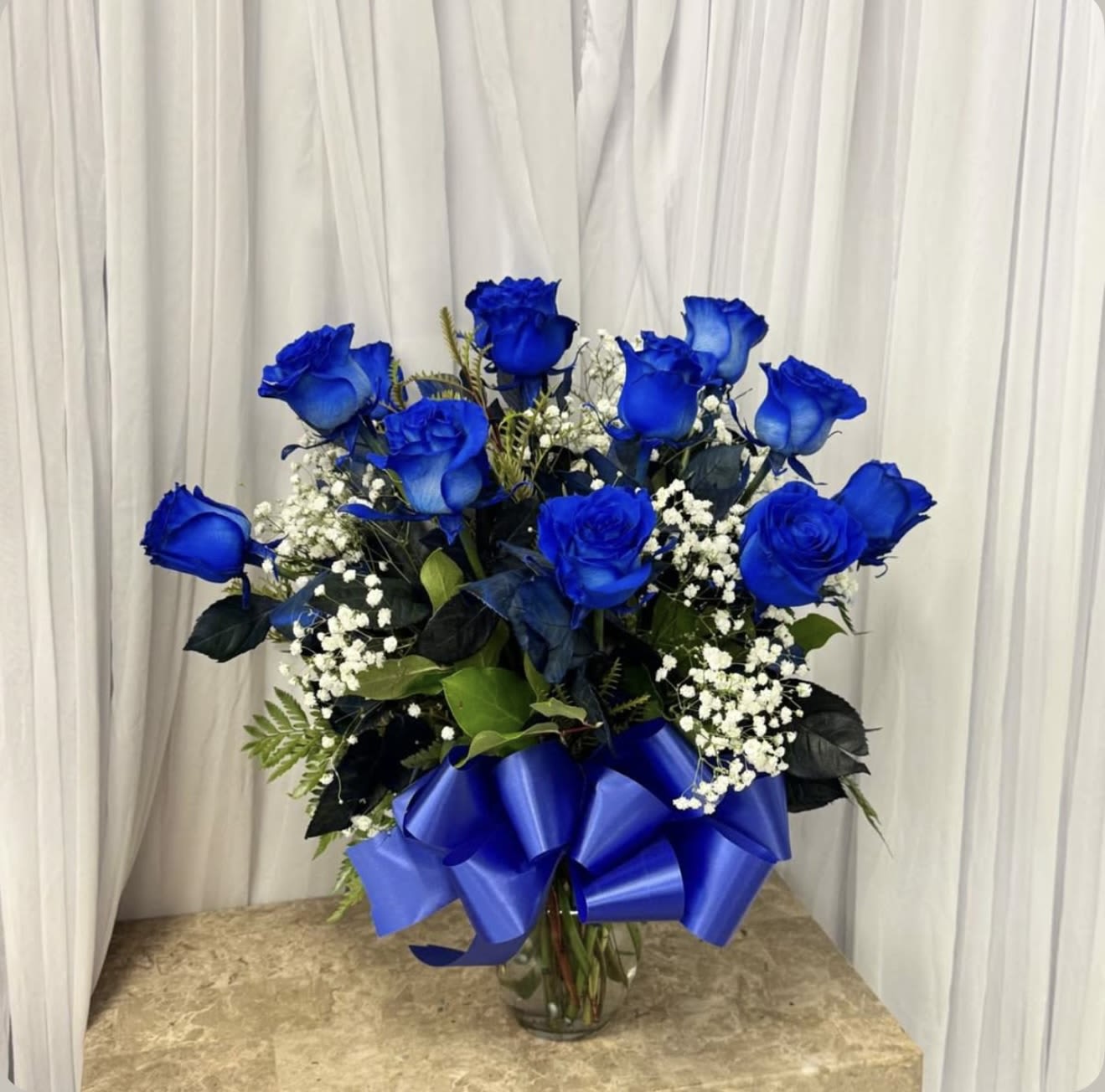 12 blue roses for him!  - Vivid cobalt-blue roses take center stage in this bold flower arrangement, a dramatic choice for an anniversary, birthday, or romantic gesture for him. The blue roses and white baby's breath create a striking contrast that feels both formal and celebratory, perfect for the person you want to honor with something unforgettable.  Tightly clustered in a rounded, upright design, twelve blue roses are softened by delicate sprays of white filler and dark green foliage for depth and texture. The stems arrive arranged in a clear glass vase and finished with a wide satin blue ribbon tied around the vase, giving the presentation a polished, monochromatic look that reads modern and intentional.  This is the kind of bouquet that makes someone feel seen-confident, special, and celebrated. Vase-arranged for easy display; refresh the water every couple of days and trim stems if you transfer to another container to extend enjoyment.