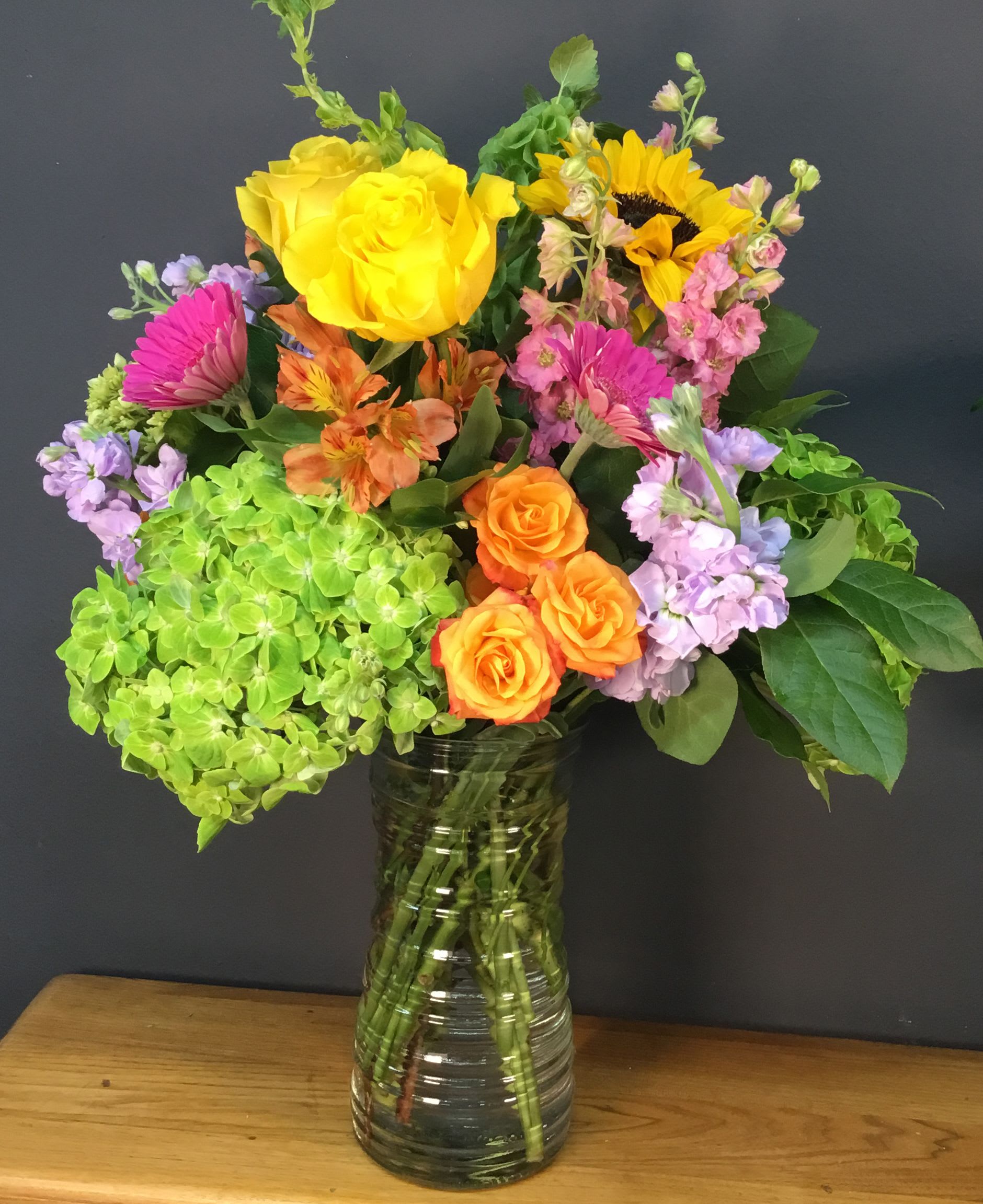 Summer Sunshine - Bright, lively color and a big cheerful sunflower center this Summer Sunshine flower arrangement - a joyful mixed bouquet perfect for birthdays, congratulations, or any moment you want to brighten someone's day. The combination of yellow roses and a single bold sunflower with pops of orange roses and alstroemeria reads sunny and celebratory; the palette naturally suits summer gatherings, a get-well pick-me-up, or to say "thinking of you" in a way that feels genuinely uplifting.