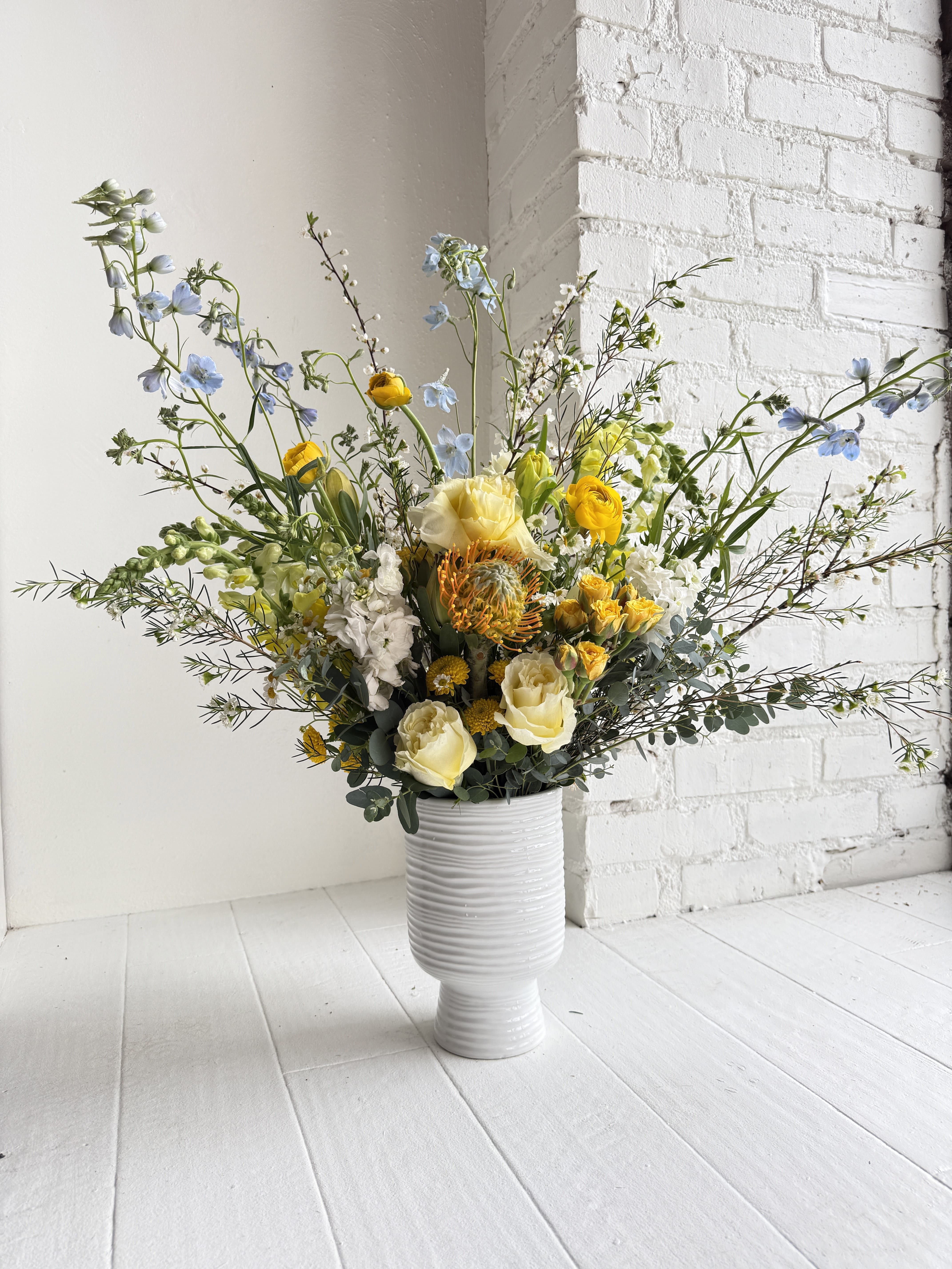 Eliza - This arrangement is a showstopper. A gorgeous mix of buttery yellows, creamy soft whites, accented with pops of blue. We carefully select our favorite seasonal flowers and artfully arrange in a tall footed vase. The overall size is approximately 32" tall, 18" wide.  We favor garden roses, ranunculus, lisinathus, heirloom mums, stock, hydrangea, snapdragons, sprirea, wax flower and other rare varieties we gather.