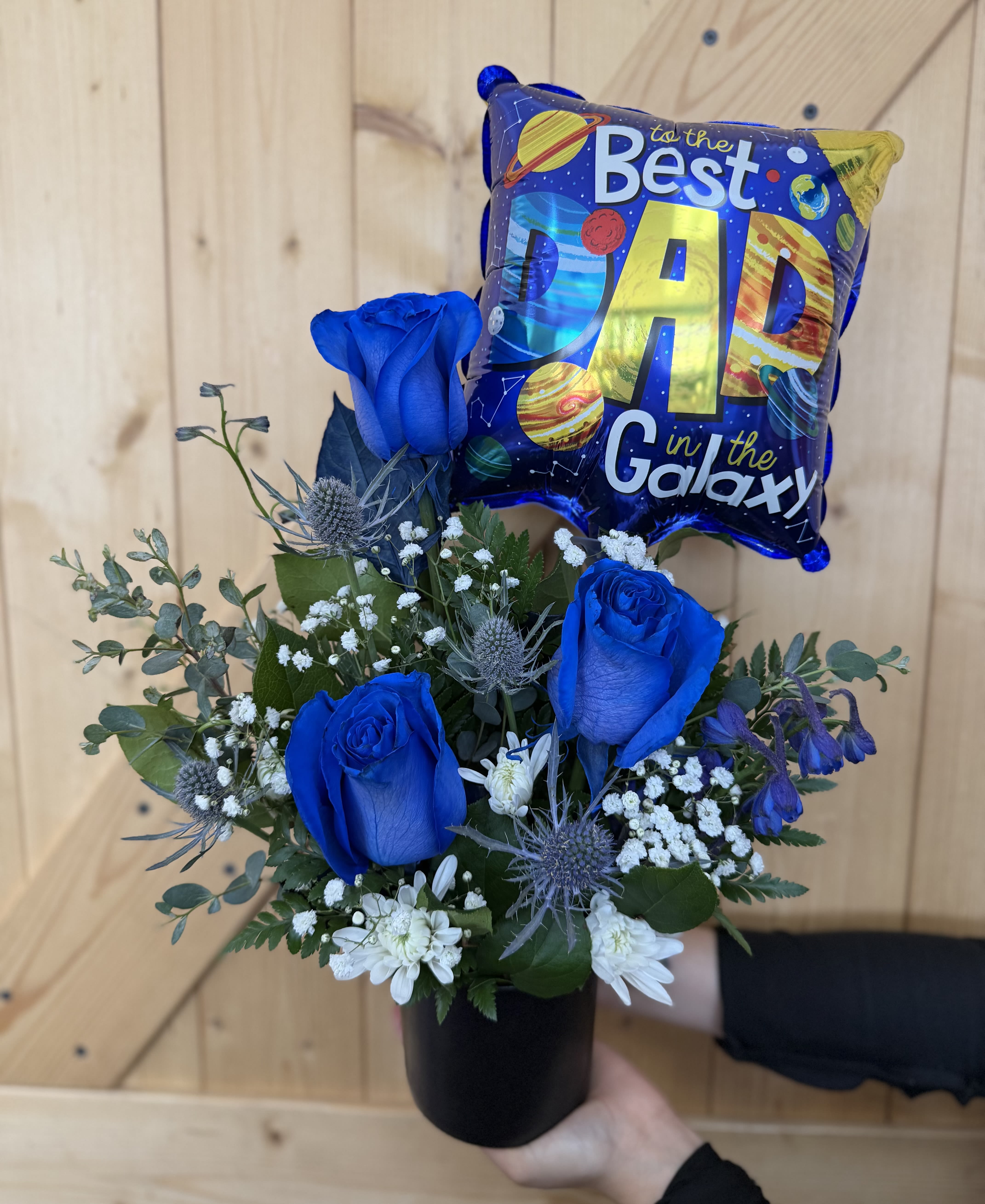 Stellar Dad - A striking Father’s Day arrangement featuring vibrant blue roses, white accent blooms, textured thistle, and lush greenery, topped with a “Best Dad in the Galaxy” balloon. Designed in a modern black container, this one sided arrangement is a thoughtful way to celebrate the dad who means the world and beyond.