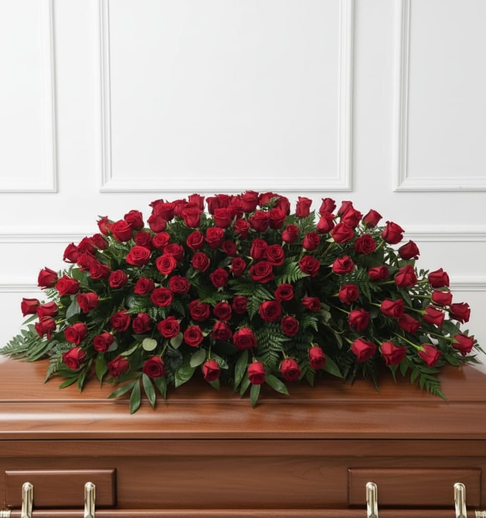 Red Tribute - A broad, low casket spray of deep red roses and lush green foliage creates a dignified, heartfelt tribute for a funeral or sympathy service. This casket arrangement is designed specifically for a wooden casket, the tightly packed roses forming a symmetrical, tabletop silhouette that reads as both formal and profoundly respectful - the kind of display that helps the person you're honoring look and feel respected in their final farewell.  The spray features 100-125 rich red roses set into dense green greenery so the flowers sit close together in a continuous, lush carpet across the casket top. Its low profile keeps the focus on the face of the casket while the deep red and green palette communicates lasting love and solemnity. Sized to span the width of a standard wooden casket, this composition emphasizes balance and full coverage for an elegant, cohesive presentation.  For families arranging services, this spray offers a meaningful way to convey sympathy and love at a funeral viewing or memorial service. Please let us know the delivery time when ordering so we can ensure proper fresh placement for the service.