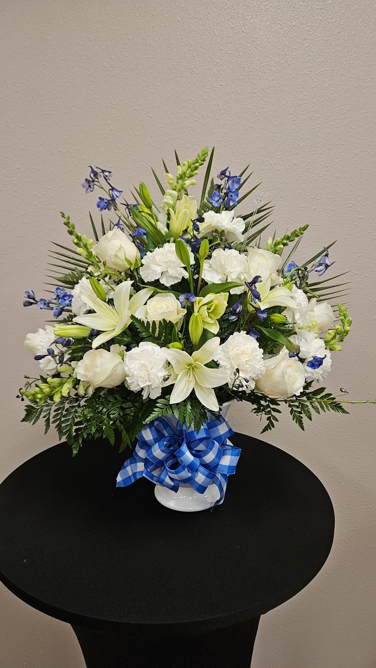 Blue &amp; White Floor Spray  - A calm, elegant floor spray in white and blue that brings a serene, formal presence to sympathy services, memorials, or any moment you want to honor someone with quiet beauty. This flower arrangement and sympathy spray pairs classic white roses and lilies with soft white carnations so the person you're thinking of feels truly respected and comforted; the tall snapdragons lend graceful height while the blue accents add a gentle, hopeful touch.  Densely layered yet airy in silhouette, the design sits above a simple white ceramic vase and is tied with a blue plaid ribbon for a finished, tasteful look. Small blue delphinium-like blooms mingle through the composition with long snapdragon spikes for vertical movement; the palette of white, blue, and green keeps the arrangement fresh and composed. Its substantial, rounded form reads well at the front of a service or as a meaningful tribute at home.  Appropriate for sympathy, memorial services, or as a dignified celebration of life, this floor spray conveys sympathy and steady support to the recipient. The vase and ribbon are included; treat lilies and roses as you would most cut flowers-keep water fresh and trim stems if re-cutting-to maintain their presence during the days they matter most.