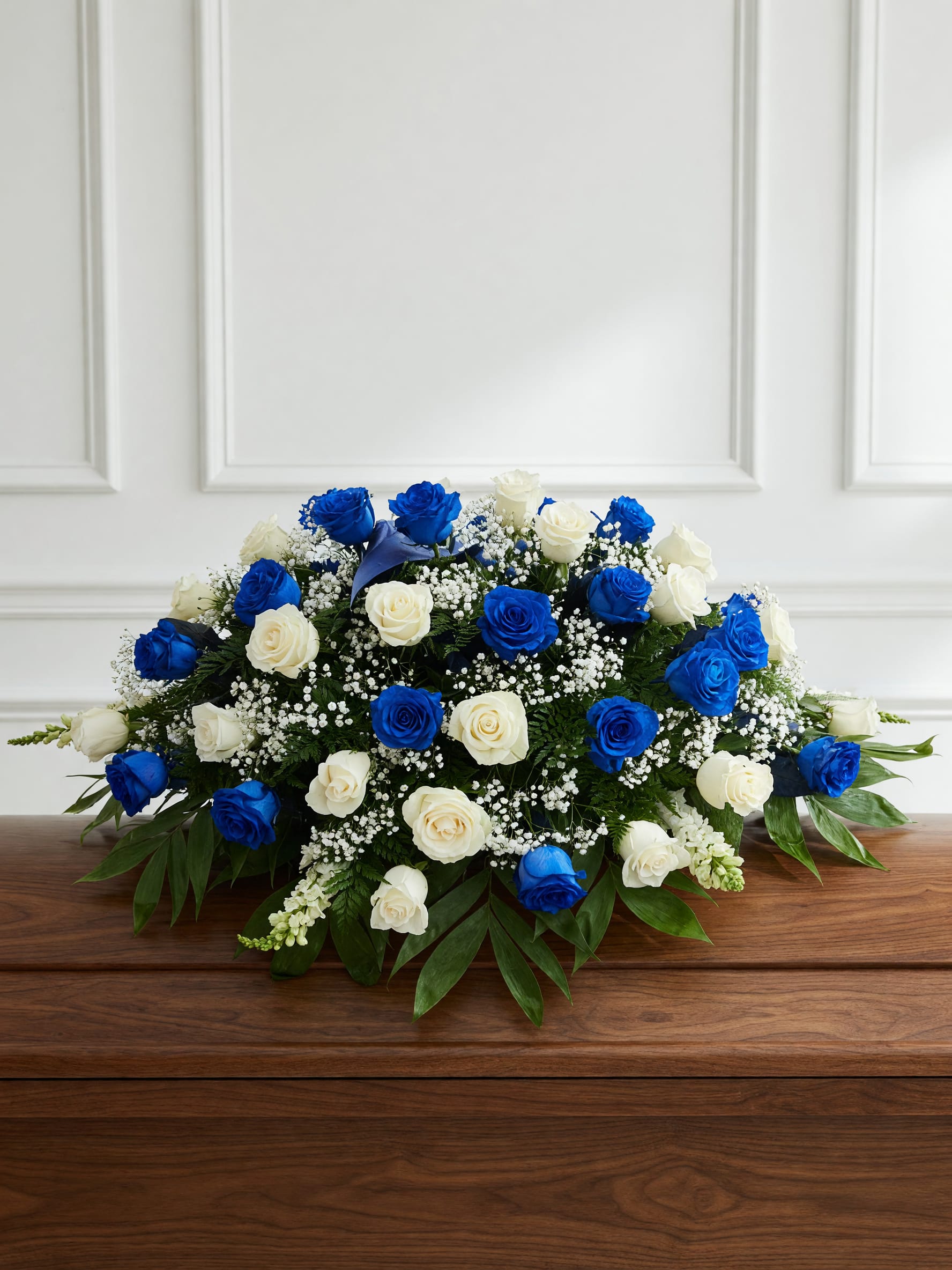 Something Blue &amp; White - A dense, casket spray of vivid blue and creamy white roses creates an elegant, serene sympathy arrangement. This flower arrangement for sympathy and memorial keeps the focus on the recipient-she'll feel comforted and seen by the calm symmetry and refined color story of blue, white, and green.  The arrangement pairs blue and white roses with clusters of tiny white filler flowers and broad glossy green foliage for a polished, formal look. Its spreading, symmetrical shape is designed to sit low and steady-perfect for placement on a casket. The contrast between the vivid blue blooms and ivory whites gives a clean, sophisticated profile.  Ideal for expressing condolences, honoring a life, or adding a touch of calm to a ceremony, this piece communicates quiet dignity and thoughtful care. Because of its low, compact silhouette it works well on tabletops and memorial stands; order with confidence knowing the recipient will receive an arrangement that reads as both graceful and respectful.