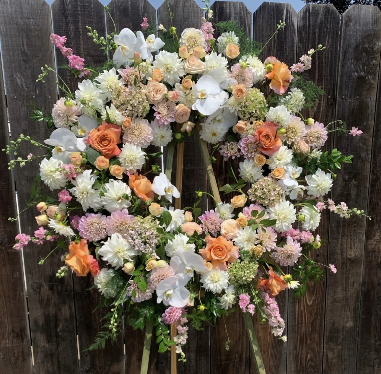 Dahlias of Grace Wreath  - A beautiful standing wreath featuring soft pastel blooms including roses, orchids, dahlias, and hydrangeas. Accented with lush greenery, this elegant arrangement is perfect for memorials or celebrations of life. Designed to offer comfort and honor with grace.