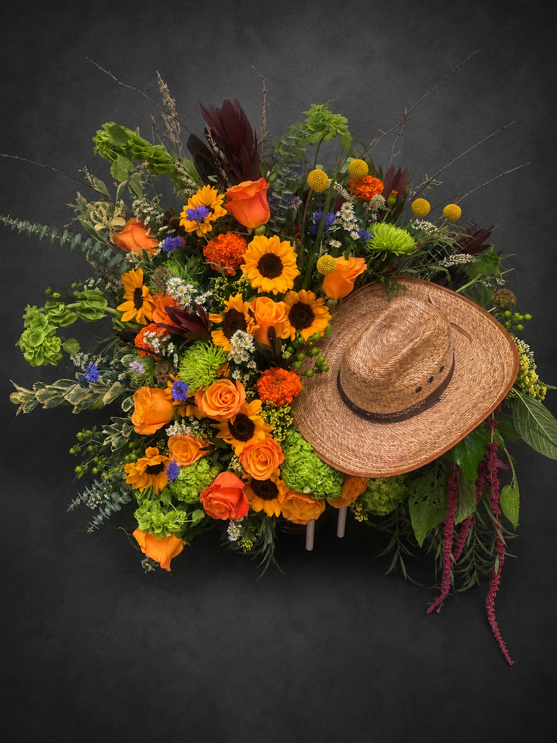Western Sunrise - A bold, country-inspired centerpiece that makes the recipient feel celebrated - this large Western Sunrise flower arrangement brings sunlit warmth and rustic charm for birthdays, congratulations, or a country-themed event. The design reads as a festive, wide-aspect bouquet and the straw cowboy hat tucked into the right side gives it unmistakable western character, so the person you're thinking of will instantly know this was chosen with personality and fun in mind.  Packed with vivid orange roses and cheerful yellow sunflowers, the arrangement also includes bright green chrysanthemums/textural blooms, yellow button-style chrysanthemums, carnations, and small daisy-like accents to create a lively, layered texture. Dark burgundy foliage and small blue accent flowers add contrast while long branching stems and trailing elements extend outward for a dramatic, expansive silhouette. The overall palette - orange, yellow, green with touches of red and mixed color accents - reads warm and autumnal yet festive year-round.  Perfect for someone who loves country flair or for celebrating milestones where bold, joyful color is right for the moment, Western Sunrise arrives as a generous, overflowing display that commands attention in any room. Because of its size and asymmetrical shape, it works well as a focal table piece or a standout gift at a party; no special care beyond fresh water is needed to keep the blooms looking vibrant in the days after delivery.