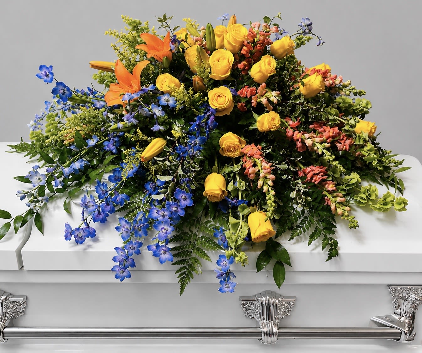 Celebration of Life - A radiant cascade of yellow roses, orange lilies, and vivid blue delphinium creates a bold funeral tribute that feels like a true celebration of life. This casket spray is designed for memorial and funeral services, offering a colorful, uplifting alternative to traditional pastels so the person you're honoring looks bright and remembered. The broad, formal presence across the casket sends a clear message of joy and respect to those gathered.  Expertly layered greenery and trailing blooms form a full, abundant shape that gently drapes over the casket, combining the softness of roses with the open, trumpet-like lilies and vertical accents of delphinium for visual movement. The arrangement's warm yellows and oranges pop against the strong blue highlights, giving both distance and depth to the composition. Sized to cover the top of a standard casket, it arrives ready to place with secure hard goods for a dignified presentation at the service.