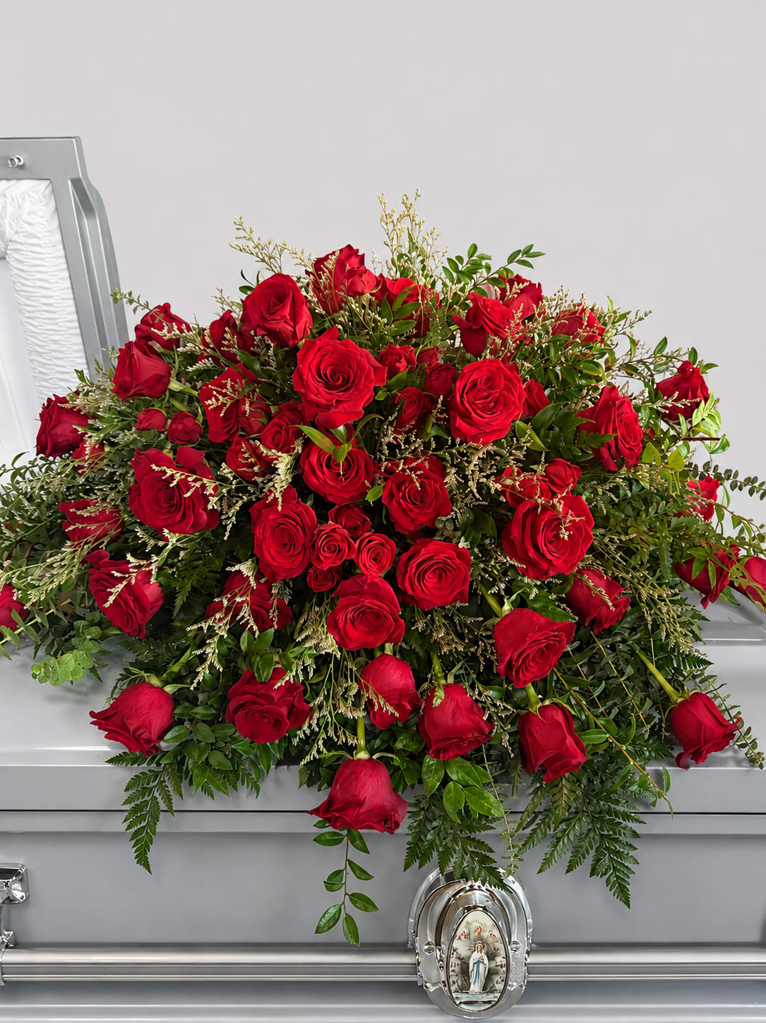 Radiant Rose Casket Spray - A generous cascade of tightly clustered red roses laid across a silver casket creates a solemn, romantic tribute ideal for a funeral or memorial service. This casket spray conveys deep love and respect; the rich red roses and lush green foliage form a traditional, formal presentation that makes the person you're honoring feel genuinely cherished.  Dense, full blooms are arranged in a long, low spray with abundant greenery and fine filler accents for texture and depth, designed to drape gracefully over a silver casket. The red-and-green contrast reads classic and reverent, while the compact cluster of roses gives a polished, handcrafted look appropriate for a loved one's final tribute.  Designed for memorial and funeral settings, this arrangement is meant to express enduring affection and sympathy at a very personal moment. Size and exact rose count can be confirmed at checkout to ensure the spray fits the casket; please contact us for delivery details, placement preferences, or any special requests.