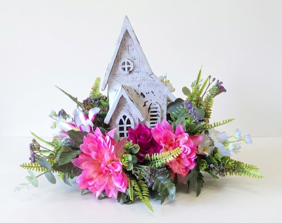Silk Wooden Church Arrangement - A charming distressed white wooden church figurine rises above a ring of silk roses, lilies, and hydrangeas to create a devotional yet cozy flower arrangement perfect for sympathy, remembrance, or a faith-centered gift. This flower arrangement for home and family occasions pairs soft peach and white with vivid and pastel pinks to make the recipient feel comforted and remembered - the kind of piece that quietly says you're holding them in your thoughts.  Soft, ruffled silk roses, lily's and dahlia blooms form a low, lush base around the wooden chapel, while two-toned pink-and-cream lilies add vertical interest and a gentle contrast in texture. The compact, low-profile design fits neatly on a side table, mantel, or entry console. 13" T 16" W