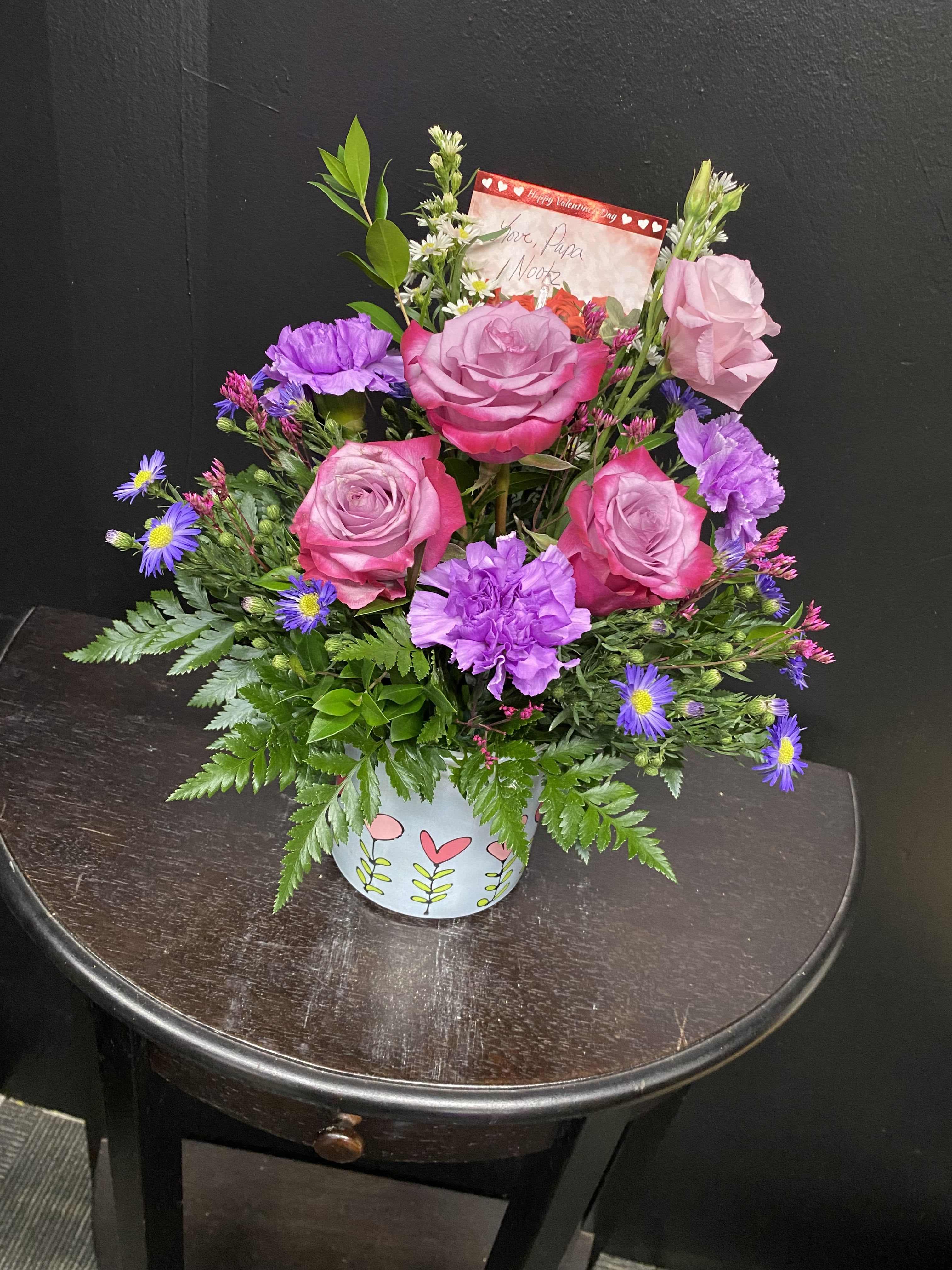 Joyful Gathering - Pink roses and lavender carnations mingle with white daisy-like asters in a bright, romantic arrangement designed as a Valentine bouquet or heartfelt anniversary gift. The cheerful palette of pink, purple, white and fresh green foliage gives the person you're thinking of a soft, affectionate impression-perfect for saying "I'm thinking of you" or celebrating a romantic moment.  This full, layered bouquet is arranged in a light blue decorative ceramic pot with a simple floral pattern for a keepsake presentation that lasts longer than wrapped stems. The mix includes several pink roses, purple carnations, small white aster filler blooms, and lush greenery for texture and volume; a matching greeting card is tucked into the back for your personal message.  Made to feel both intimate and festive, this arrangement suits romantic occasions like Valentine's Day, anniversaries, or simply to surprise a loved one. Care is simple: keep the soil lightly moist and place the pot in bright, indirect light to prolong freshness; select your size at checkout for the perfect visual impact.