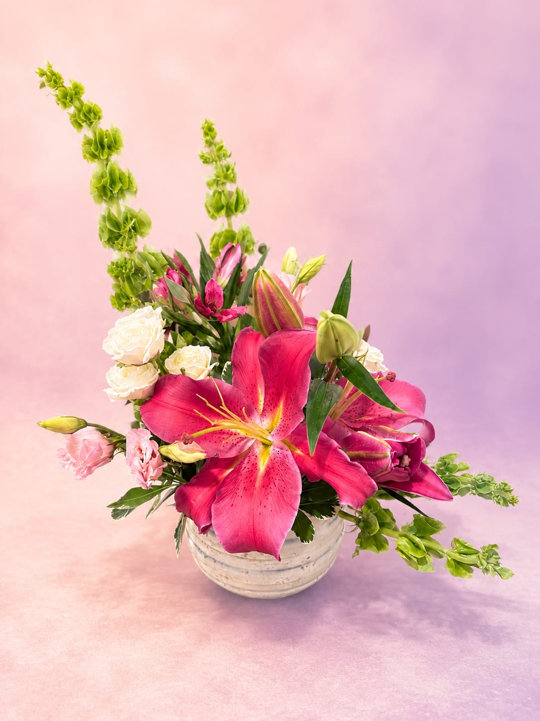 Pink Paradise - Bold, vibrant, and full of graceful movement, Pink Paradise is a stunning garden-inspired arrangement that radiates joy and elegance. Showstopping hot pink oriental lilies take center stage, surrounded by soft blush Lisianthus, creamy white roses, and vivid alstroemeria. Fresh green bells of Ireland add height and whimsical charm, creating a design that feels both lush and artistic. Arranged in a textured ceramic keepsake bowl, this arrangement makes a beautiful statement for birthdays, anniversaries, celebrations, or simply brightening someone’s day.