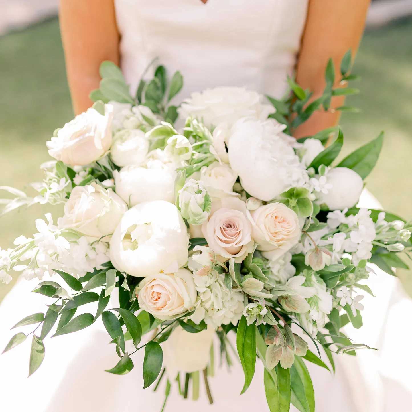 Blush Bouquet - The lush, garden-style, hand-tied bouquet, features pillowy white peonies, elegant garden roses, white blossoms, ranunculus buds, delicate clusters of white stock, lilac blossoms, and unique ruffled petals, giving off a timeless romantic feel.