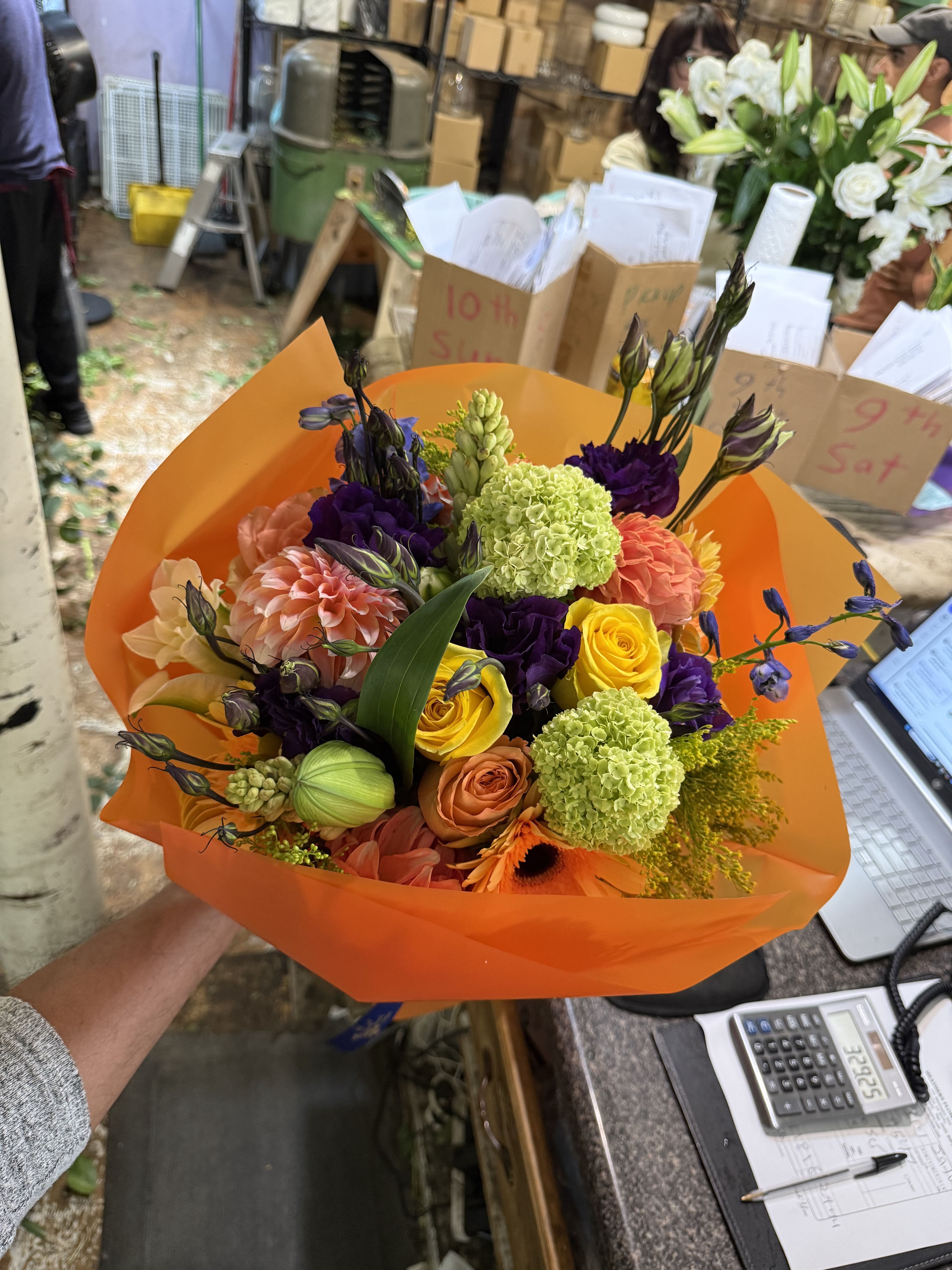 Citrus Symphony  - This bouquet is a vibrant celebration of color and texture, artfully wrapped in bold orange paper that perfectly complements its warm, cheerful palette. Featuring sunny yellow roses, peach dahlias, orange gerbera daisies, lime green viburnum, and rich purple lisianthus, it strikes a beautiful balance between balance between elegance and playfulness. The combination of bright citrus tones, soft blush hues, and deep violet accents creates a bouquet that feels energetic, modern, and full of life. 