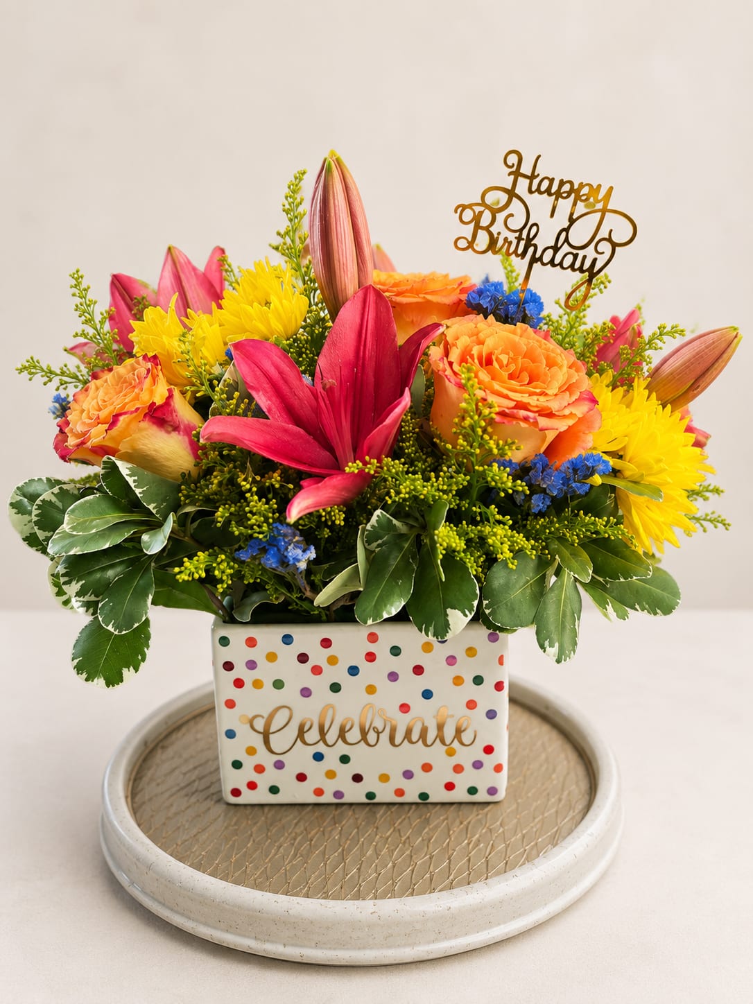 Birthday celebration garden - vibrant birthday arrangement featuring bright pink lilies, peach roses, yellow chrysanthemums, blue accent flowers, and premium greenery arranged in a festive keepsake ceramic container. Finished with a gold "Happy Birthday" topper, this cheerful design is perfect for celebrating someone's special day with color and elegance.