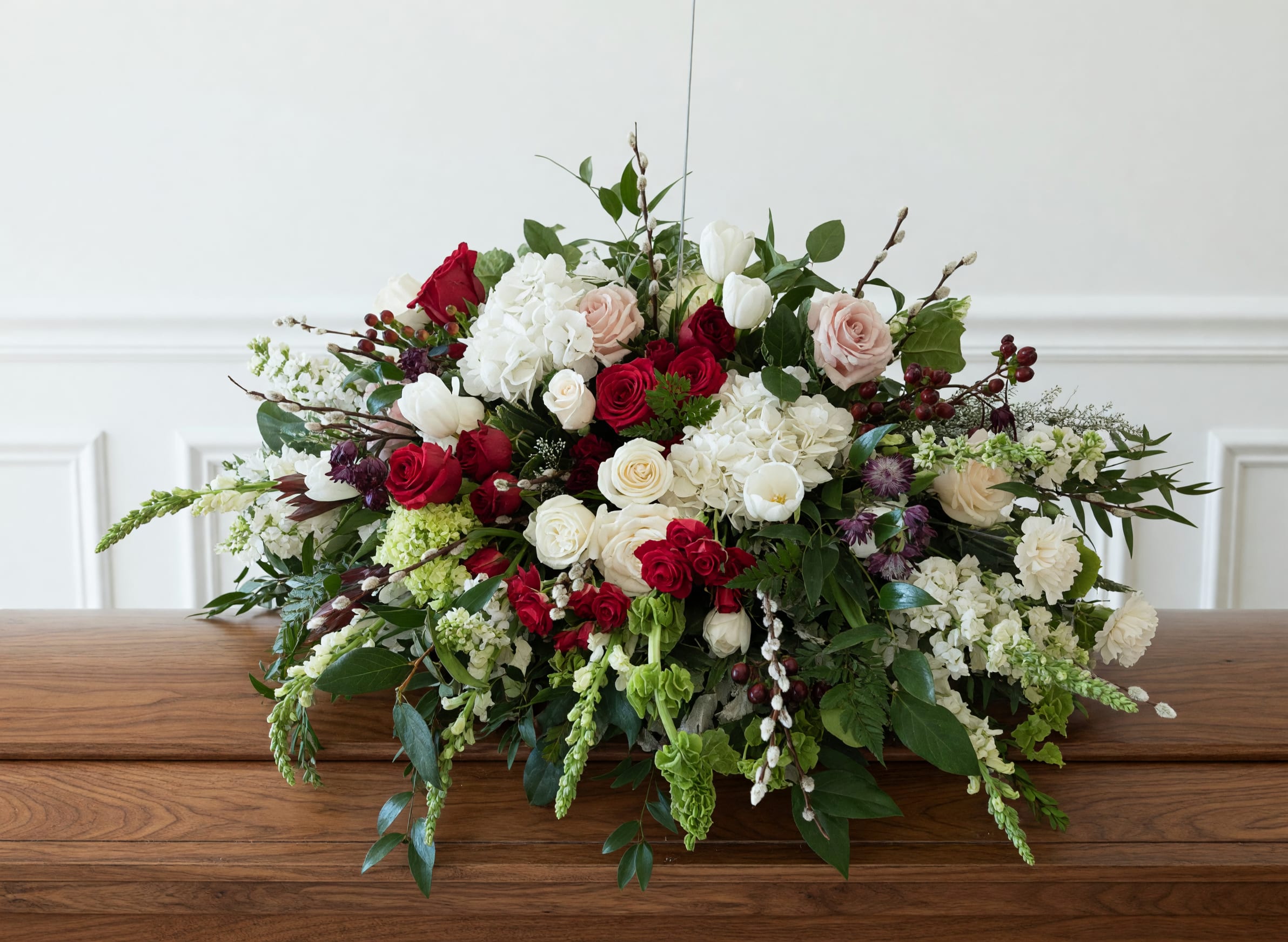 Crimson and Ivory casket cover  - This abundant casket spray features velvet red roses and seasonal blooms in whites, creams and ivory shades. This elegant tribute conveys love, respect and everlasting remembrance.  Please call the shop directly to discuss color palettes or specific flowers.  Approx 48"L x 30" H 