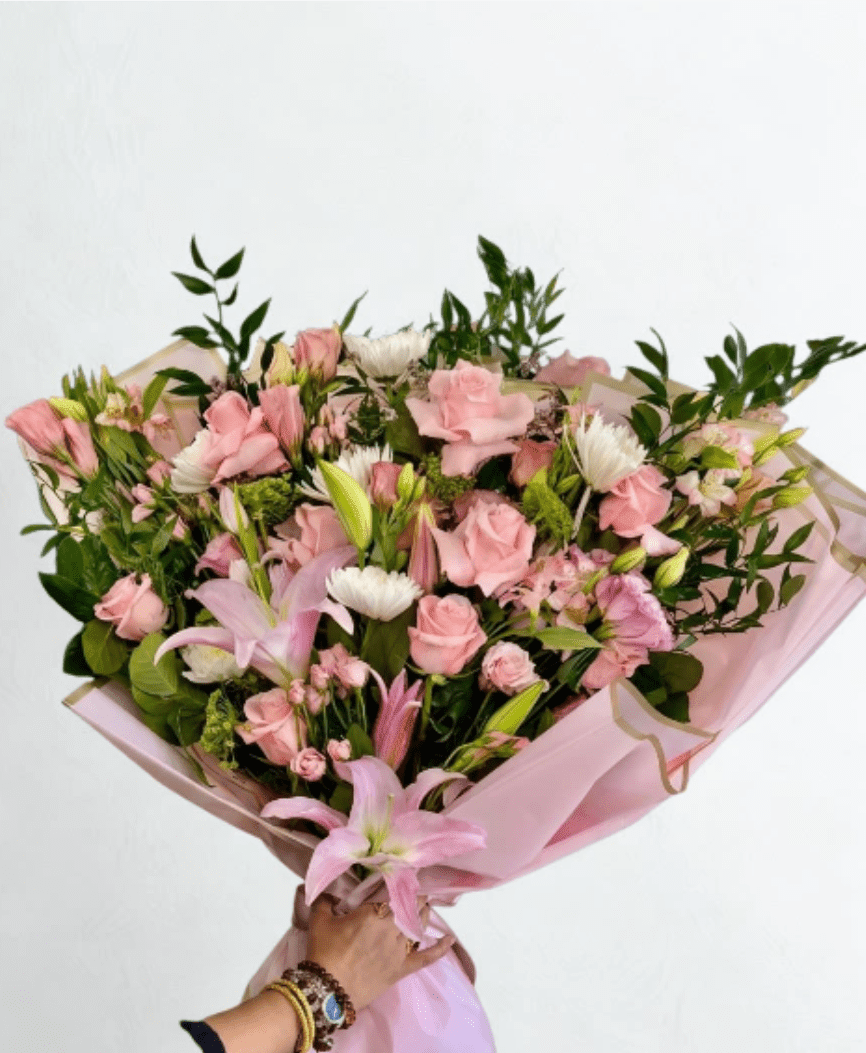 The Regal Romance by The Flower Spot™ - A lavish, hand-tied bouquet that reads like a love letter in soft pastels-perfect as a birthday bouquet, anniversary roses alternative, or a thoughtful "thinking of you" arrangement. The recipient will feel genuinely celebrated the moment they see the lush mix of pink roses and stately lilies framed by white daisy-like blooms, lavender filler, and fresh green accents-a romantic presentation that says affection and elegance.  Carefully curated by our designer, this large arrangement layers open blush roses, lily buds for height, chrysanthemums, lisianthus, alstroemeria, and airy filler florals to create a full, textured silhouette. Pale pink wrapping paper and a dual ribbon-satin with a gold-edged trim-give it a polished, gift-ready finish. The bouquet arrives densely packed and wide-spread for an abundant look; expect a generous, hand-tied size that makes a striking table centerpiece or focal gift.  Ideal for anniversaries, Mother's Day, birthdays, or simply to make someone feel truly seen, this arrangement is arranged to hold water well until it's transferred to a vase. For best longevity, recut stems, change water daily, and keep away from direct heat-ordering is available for same-week local delivery and careful gift wrapping ensures it arrives ready to impress.