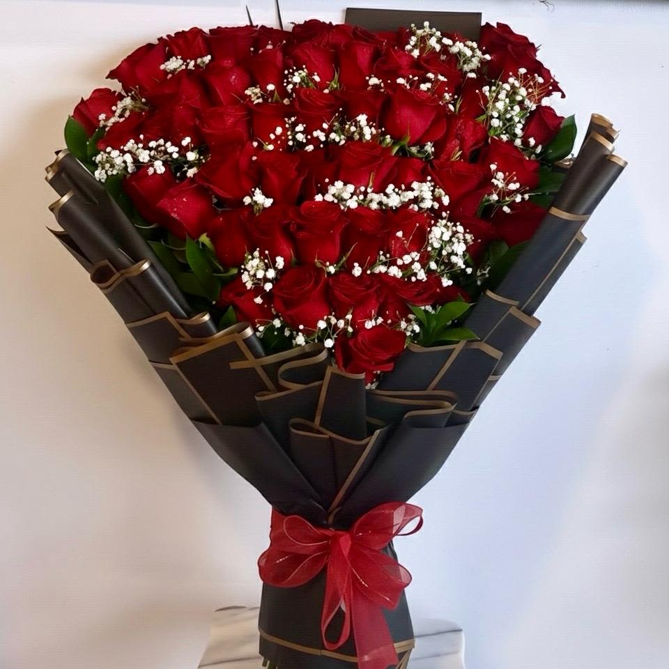 Infinite Love by The Flower Spot™ - A dense cluster of fifty premium red roses arranged with delicate white baby's breath creates an immediate, romantic impact-perfect as an anniversary bouquet or a bold birthday surprise. The dramatic red-and-black contrast and hand-tied presentation make the recipient feel deeply celebrated; this is the kind of arrangement that reads as a grand, personal declaration of love or appreciation.  Each rose is carefully curated and gathered into layered black wrapping paper with subtle gold trim, then finished with a sheer red ribbon for a luxe, tactile finish. The compact, couture-style bunch emphasizes rich red petals against crisp white accents and verdant green foliage, giving it a classic yet modern silhouette that photographs beautifully and stands out in person.  Ideal for anniversaries, Valentine's Day, Women's Day, or milestone birthdays, this bouquet arrives ready to display-no extra fuss required. To keep the roses looking their best, trim stems, place in fresh water, and change water daily; built for dramatic gifting, it's the kind of present that makes her feel truly seen.