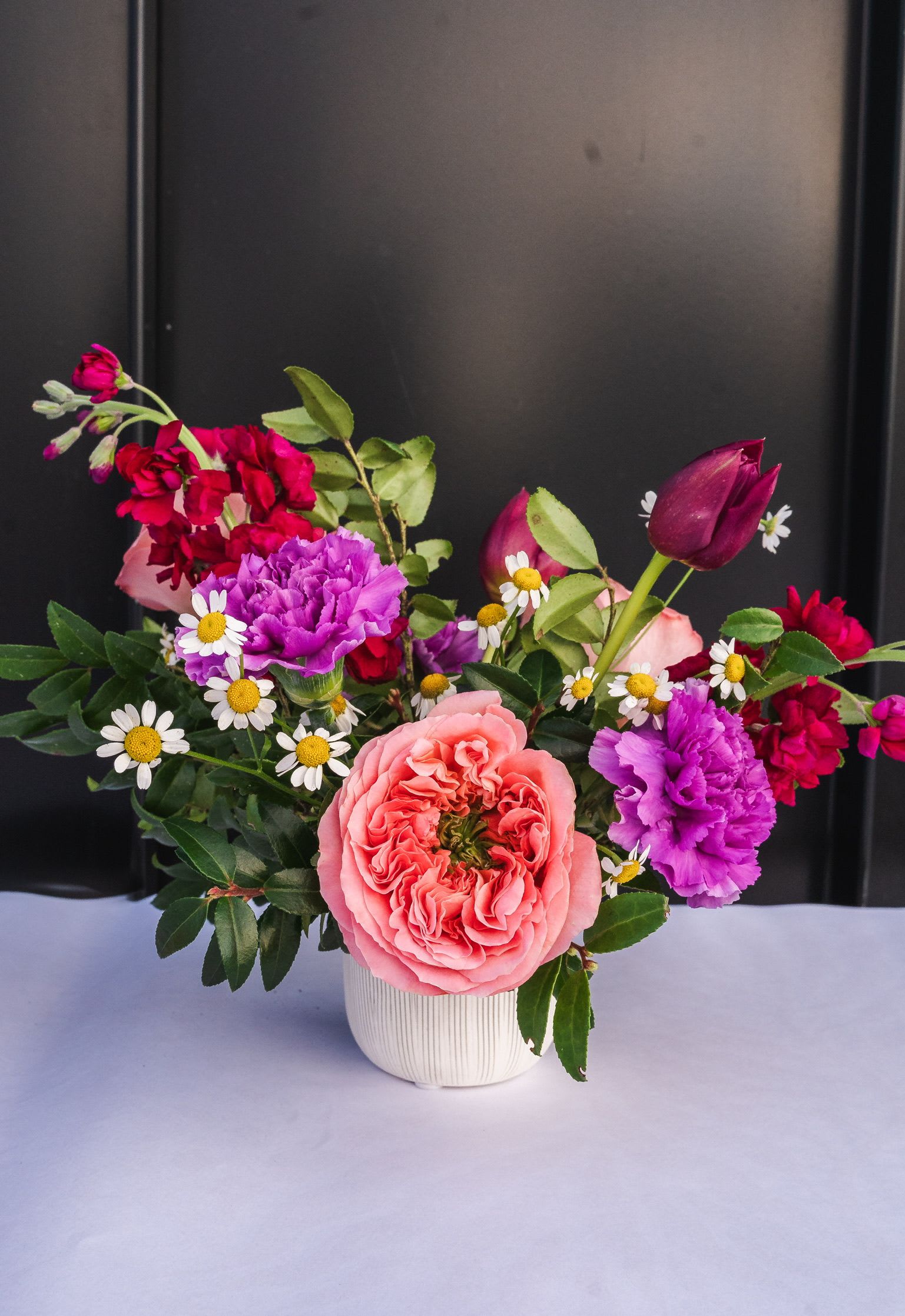 Compact arrangement of pink roses, purple carnations, red stock, and small white daisies in a white ceramic vase.