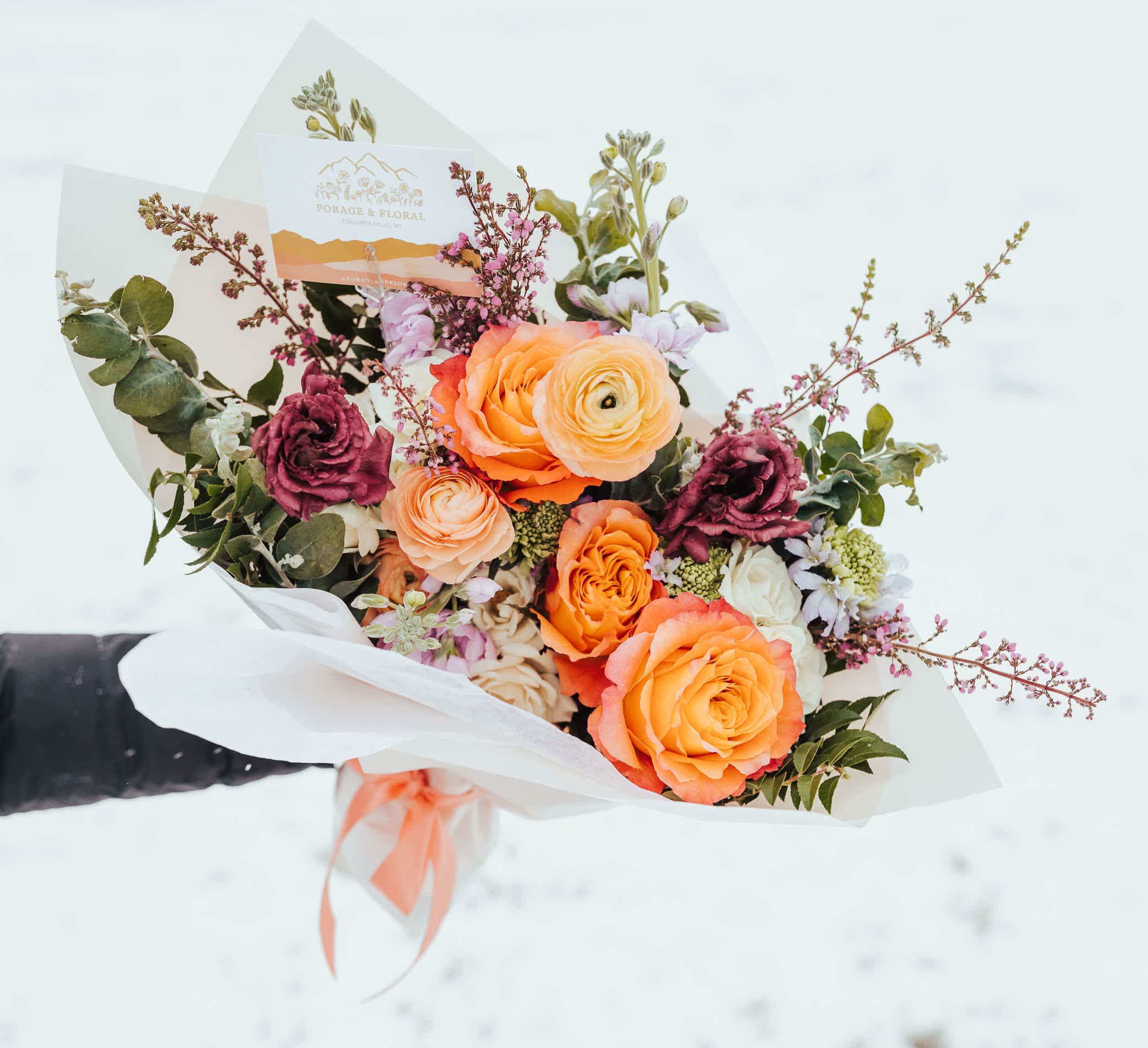 Hand-tied bouquet of orange and yellow roses and ranunculus with burgundy blooms in white wrap