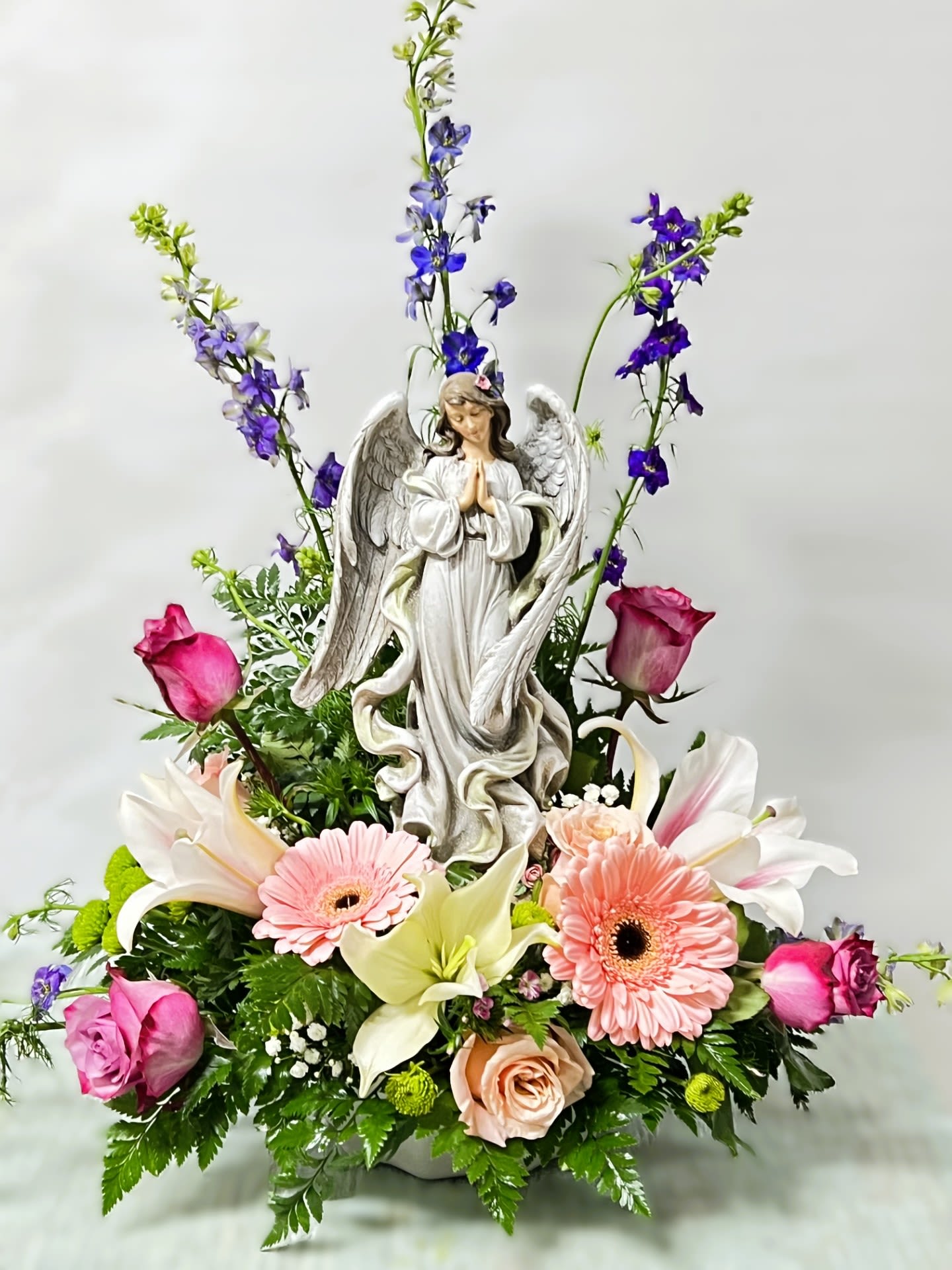 Angel figurine surrounded by pink roses, lilies, gerberas, and purple flowers