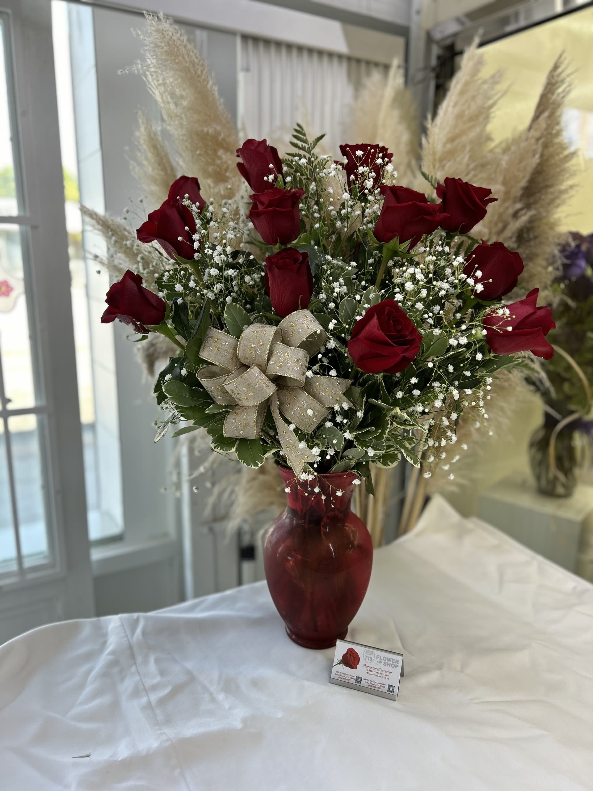 Tall arrangement of red roses with white filler flowers in a red glass vase with a gold bow