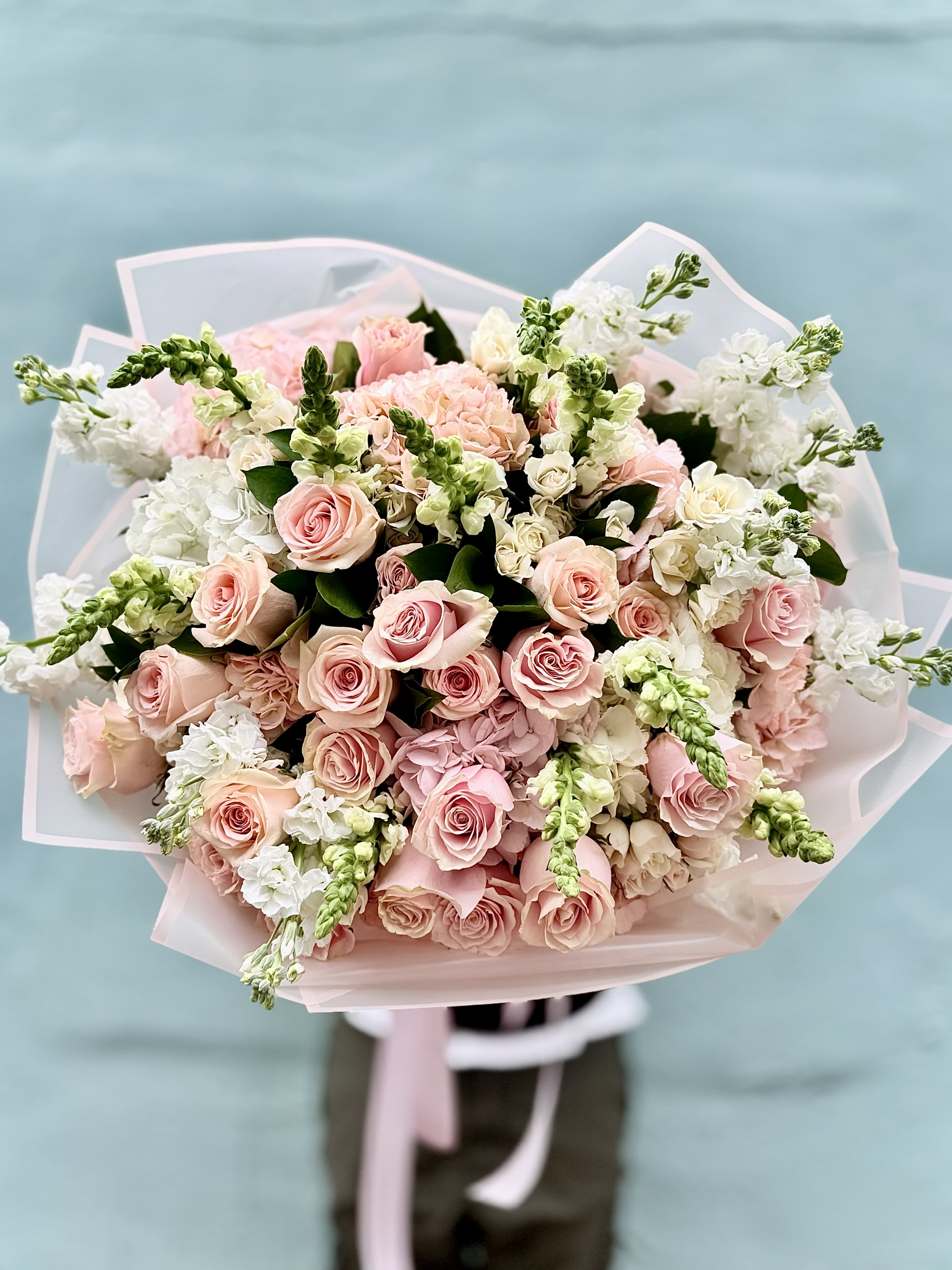 Pastel Dream - A cloud of pale pink roses takes center stage in this hand-tied flower arrangement, immediately reading as a romantic birthday bouquet, anniversary gift, or Mother's Day surprise. The soft blush roses are complemented by airy white stock and hydrangea, with green snapdragon spires and foliage adding vertical texture-together they create a gentle, garden-fresh impression that makes the recipient feel truly celebrated.  Closely packed for a lush, rounded silhouette, the mix of tightly opened rose heads and delicate white spikes gives depth and a refined, layered look. The bouquet is finished in translucent and soft pink wrapping paper with a matching pale ribbon for a polished, gift-ready presentation; it can also be delivered in a clear vase upon request. Ideal for thank-you gifts, spring celebrations, or quiet romantic gestures, Pastel Dream arrives ready to brighten a room and linger as a thoughtful, elegant reminder that someone is being thought of.