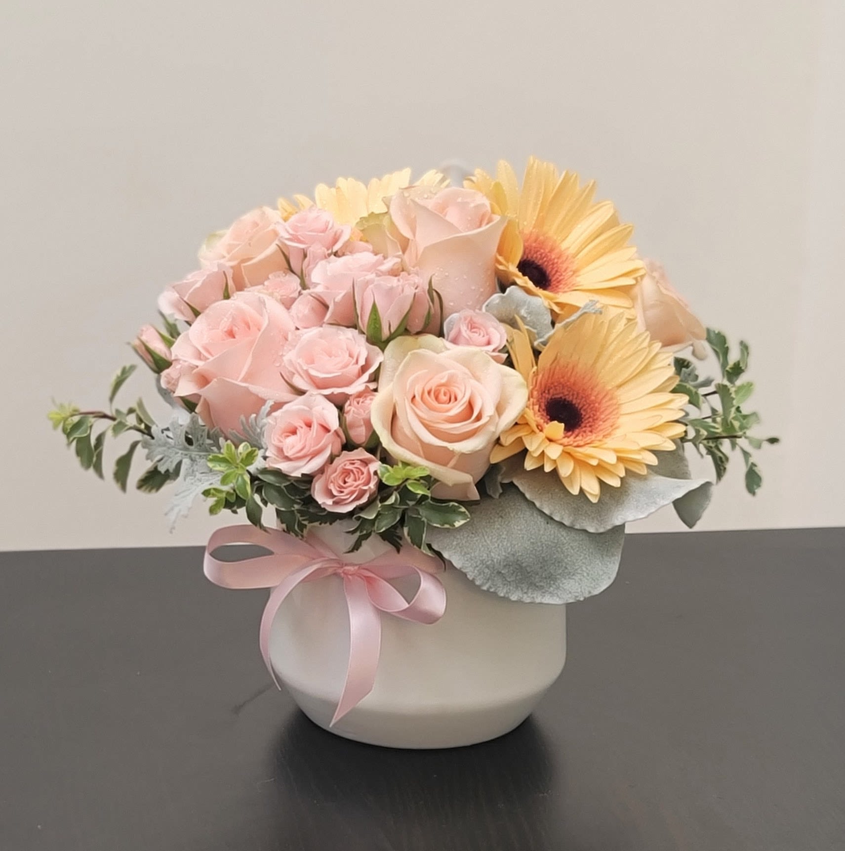 Pink roses and yellow gerbera daisies in a white vase with a pink ribbon
