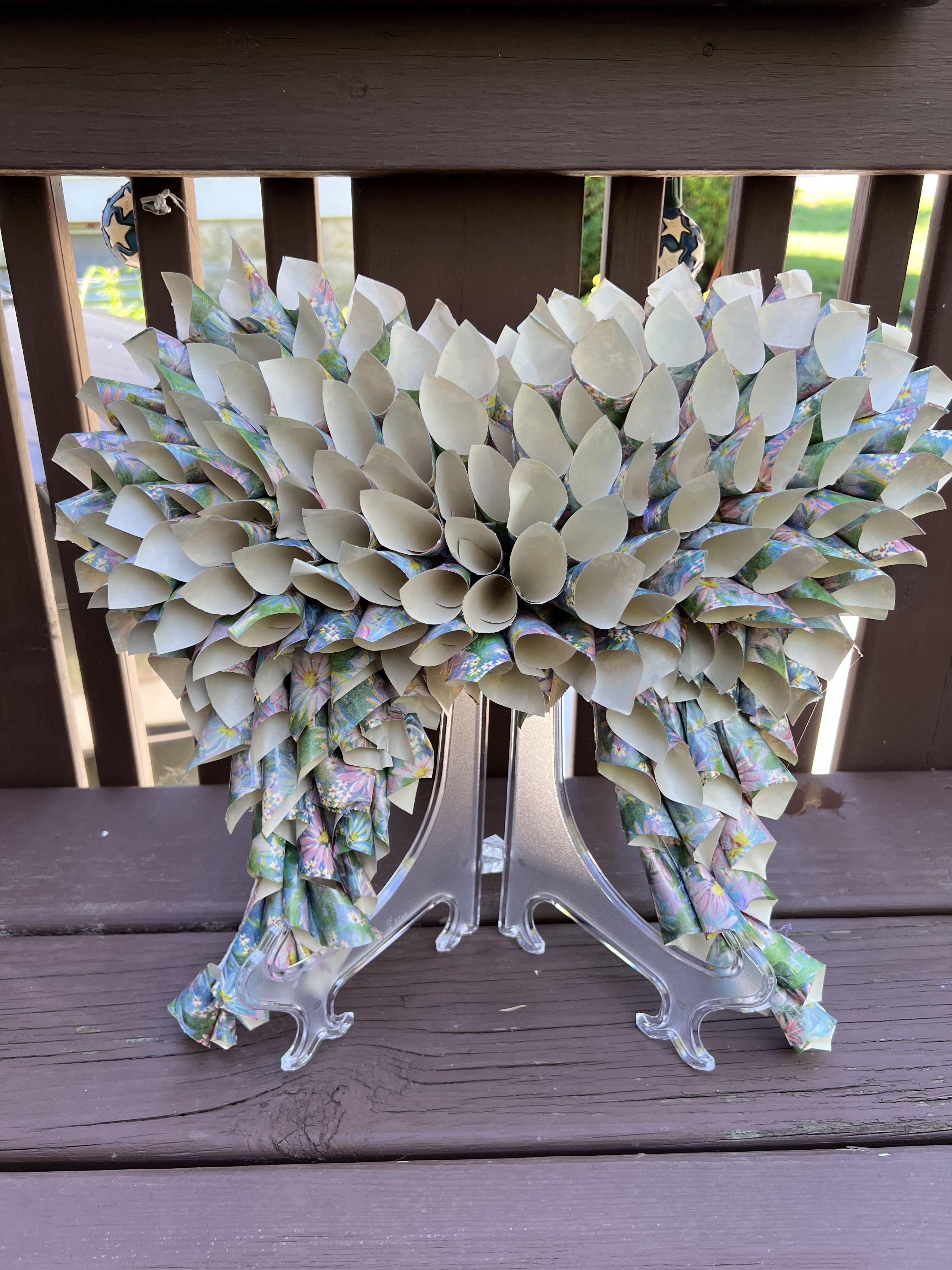 Angel Wings - Angel Wings is a large (12 X 12) decorative paper flower arrangement that reads like a handcrafted keepsake - perfect as a celebratory centerpiece or a lasting gift for the service. The airy white petals and soft pastel floral-patterned paper create a bright, uplifting presence that will make the person you're thinking of feel cherished and noticed, without the need for fresh-flower maintenance.  Made from tightly rolled white paper petals with hints of mixed-color, pastel floral print woven throughout, this arrangement spreads broadly for a full, textured look. It sits on a clear display stand so it presents beautifully on a mantel, entry table, or as an event focal point; the paper wrapping and finished base give it a polished, crafty charm that reads equally as a long-term decorative accent.  Because it's a keepsake-style arrangement, Angel Wings requires no watering and keeps its shape and color indoors away from direct prolonged sunlight to preserve the paper. Choose this when you want a cheerful, non-traditional floral gift that will continue to delight long after the occasion.