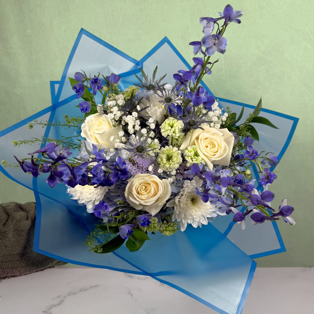 Whispering Winds Wrapped Bouquet - The Whispering Winds Wrapped Bouquet by Savilles Florist is a magnificent display of blooms, sure to wow anyone who receives one! A mix of stunning blues and whites are combined in a delicate blue wrap, tied with a sheer blue ribbon, ready to be handed off to someone as a wonderful gift. Flowers include white roses, blue hydrangea, blue delphinium, and more, along with lovely filler like penny cress and statice.