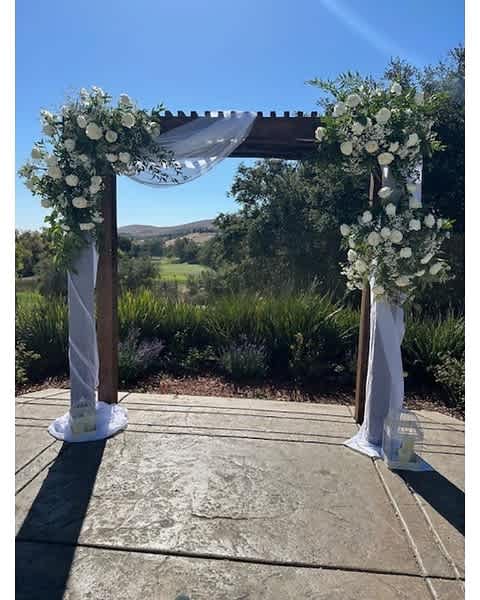 Outdoor wooden arch with white floral arrangements and sheer white draping fabric