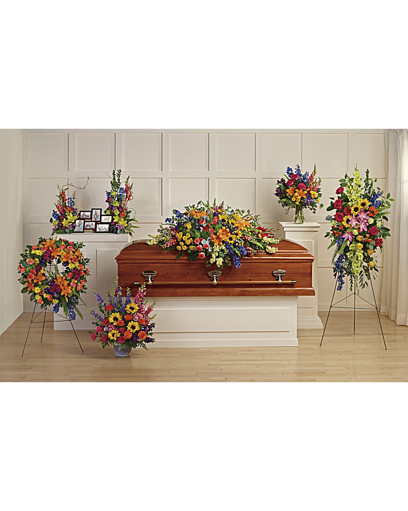 Colorful funeral floral set with casket spray, standing sprays, wreath, and vase arrangements around a wooden casket.