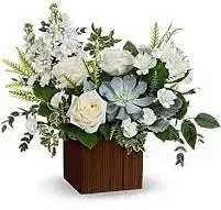 Just Breathe - A soft, cool-toned arrangement that immediately soothes-Just Breathe is a white and green floral design with a pale blue focal bloom that draws the eye and calms the room. As a sympathy arrangement, thank-you gift, or simple thinking-of-you bouquet, this flower arrangement offers a quiet, comforting presence that makes the person you're thinking of feel seen and supported.  This composition pairs white roses, creamy hydrangeas, white daisies and delicate filler blooms with varied greenery for texture, all gathered tight for a full, layered look. The pale blue flower at the center provides a gentle contrast against the rounded petals and airy accents, while the square wooden cube vase adds a modern-rustic touch that sits well on a mantel, bedside table, or office desk.  Ideal for sending comfort, gratitude, or a peaceful hello, Just Breathe is sized to make an everyday statement without overwhelming a small space. Arrange for local delivery to ensure the blooms arrive fresh; misting the flowers and keeping them out of direct heat will help extend their display.