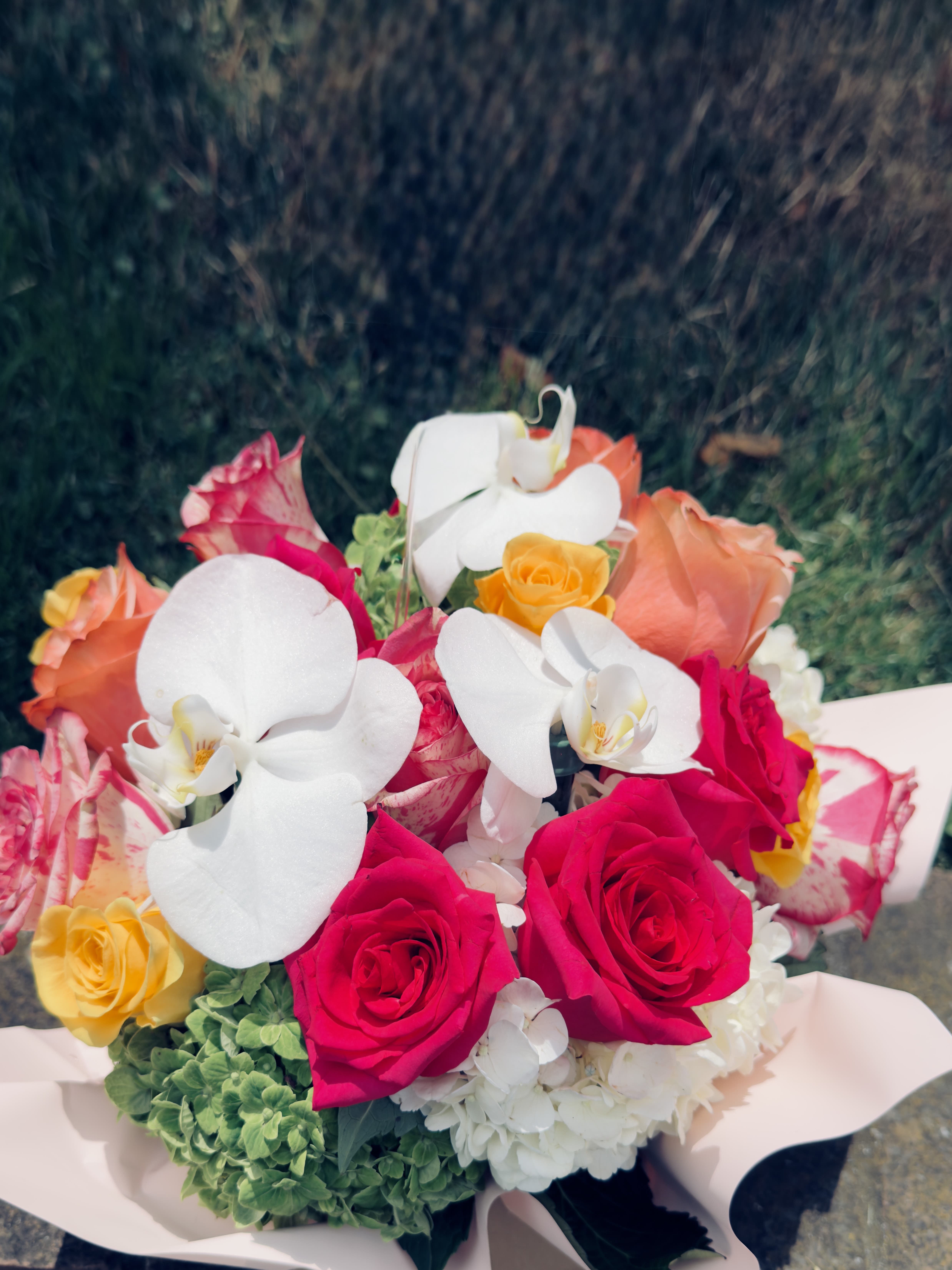 Rose orchid arrangment - A vivid mix of hot pink, coral and bright yellow roses paired with sculptural white Phalaenopsis orchids makes this flower arrangement an unforgettable birthday bouquet, anniversary surprise, or romantic gift. The contrast of saturated roses against the clean, elegant orchids creates a lively, romantic impression that will make the person you're thinking of feel truly celebrated.  Generous white hydrangea at the base gives the arrangement volume and a soft cloud of texture, while the pale pink bouquet wrap keeps the presentation polished and gift-ready. Colors include pink, white, yellow and mixed tones; flowers are roses, Phalaenopsis orchids, and hydrangeas, hand-tied and finished in a bouquet wrap for easy gifting.  Perfect for marking milestones or simply saying "you're special," this arrangement reads as both joyful and refined-ideal for a partner, close friend, or mother who appreciates bold color with elegant accents. Order with standard sizing; care is simple-keep flowers in fresh water and away from direct heat to enjoy their beauty as long as possible.