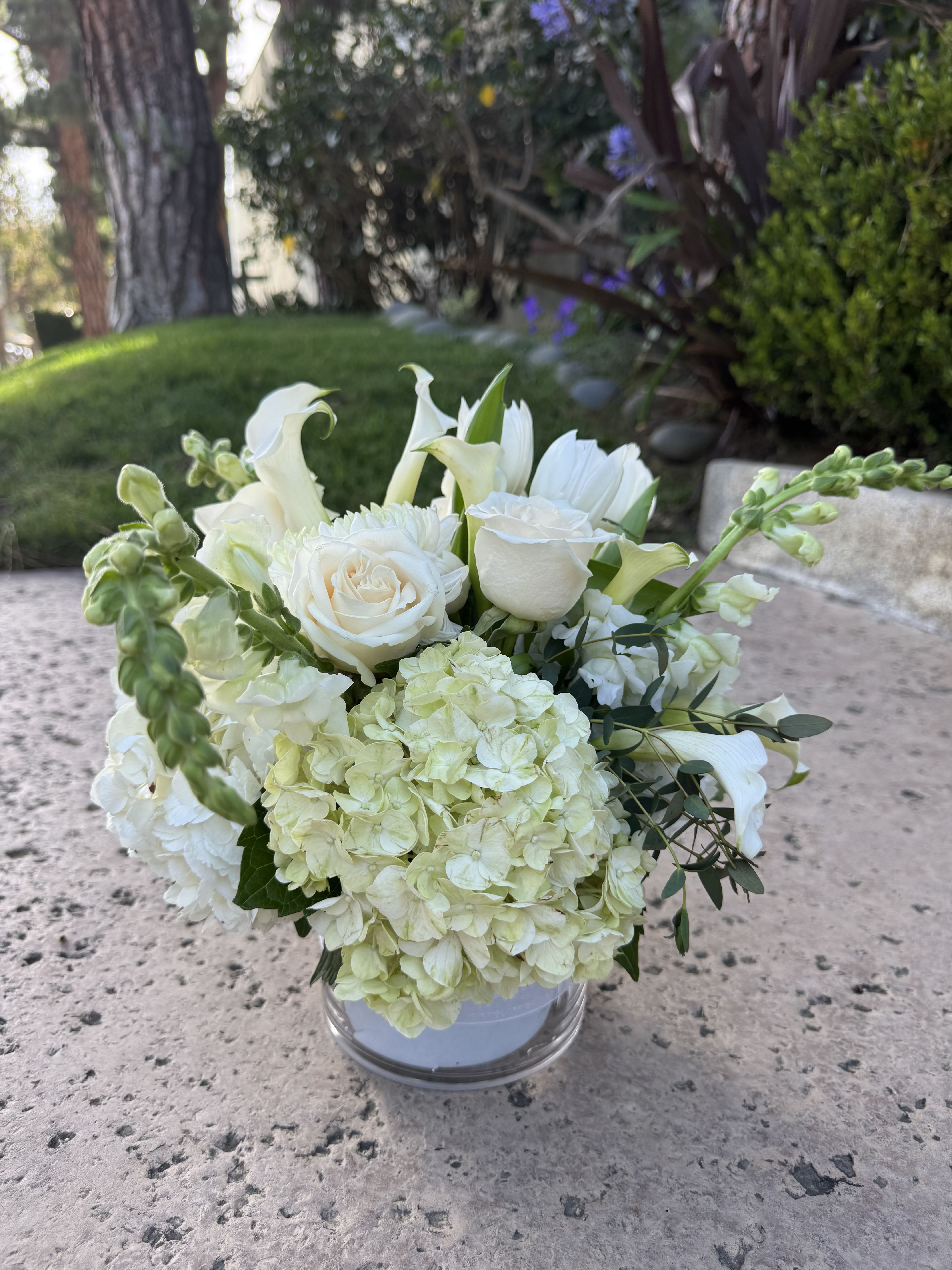 Hydrangea, rose arrangment  - A compact, rounded bouquet of white roses, pale hydrangea, and graceful white calla lilies creates a serene centerpiece perfect for a wedding bouquet or a sympathy arrangement. The clean white and soft green palette reads formal and comforting, the kind of floral gift that makes the person you're thinking of feel soothed and truly honored.  Densely layered blooms sit in a clear glass vase so petals and stems form the visual focus; soft green foliage and a few trailing stems add gentle movement and texture against the smooth white flowers. The arrangement's compact shape gives it a polished, table-ready presence while the mix of roses, hydrangeas, and calla lilies brings a variety of forms-rounded heads, delicate trumpets, and lush clusters-for tactile interest.  Ideal for ceremonies, memorials, or to convey quiet sympathy, this arrangement sends a message of calm support and refined beauty. Standard vase dimensions and care: keep water fresh, recut stems every few days, and place away from direct heat or harsh sunlight to preserve bloom life.
