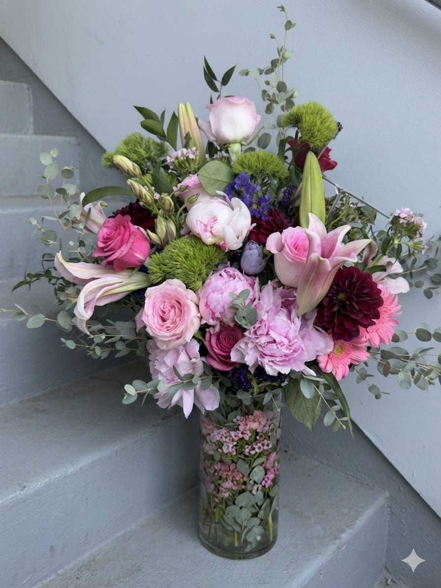 Peony, rose  Lily arrangement - A lush cascade of soft pink peonies and creamy roses meets rich burgundy dahlias and purple accents in this garden-style flower arrangement-perfect as an anniversary bouquet, wedding centerpiece, or sympathy arrangement that feels both celebratory and deeply comforting. The mix of lilies, gerbera-like daisies, and carnations gives the design a layered, romantic texture that reads like a hand-gathered bouquet arranged in a clear glass vase.  This large, full composition is presented in a clear cylindrical glass vase and features airy eucalyptus woven throughout for movement and a fresh green contrast. Expect a generous blend of roses, lilies, peonies, dahlias, carnations, and daisy-like blooms in pink, white, purple, and green tones; the burgundy accents add depth and richness to the palette. The overall effect is loose yet polished, with blooms arranged at varying heights for a natural, garden-picked silhouette.  Made to make the recipient feel truly seen, this arrangement suits milestone celebrations, intimate ceremonies, or moments when you want to offer heartfelt sympathy. The clear vase makes it gift-ready; light maintenance-fresh water and a cool spot-will help the flowers stay vibrant through the week.